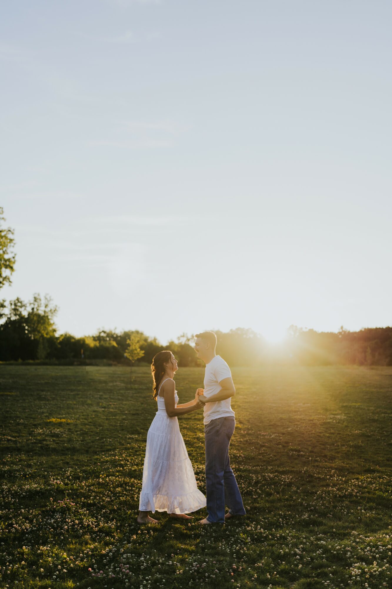 A couple holding hands in a field during sunset, with trees and a clear sky in the background.