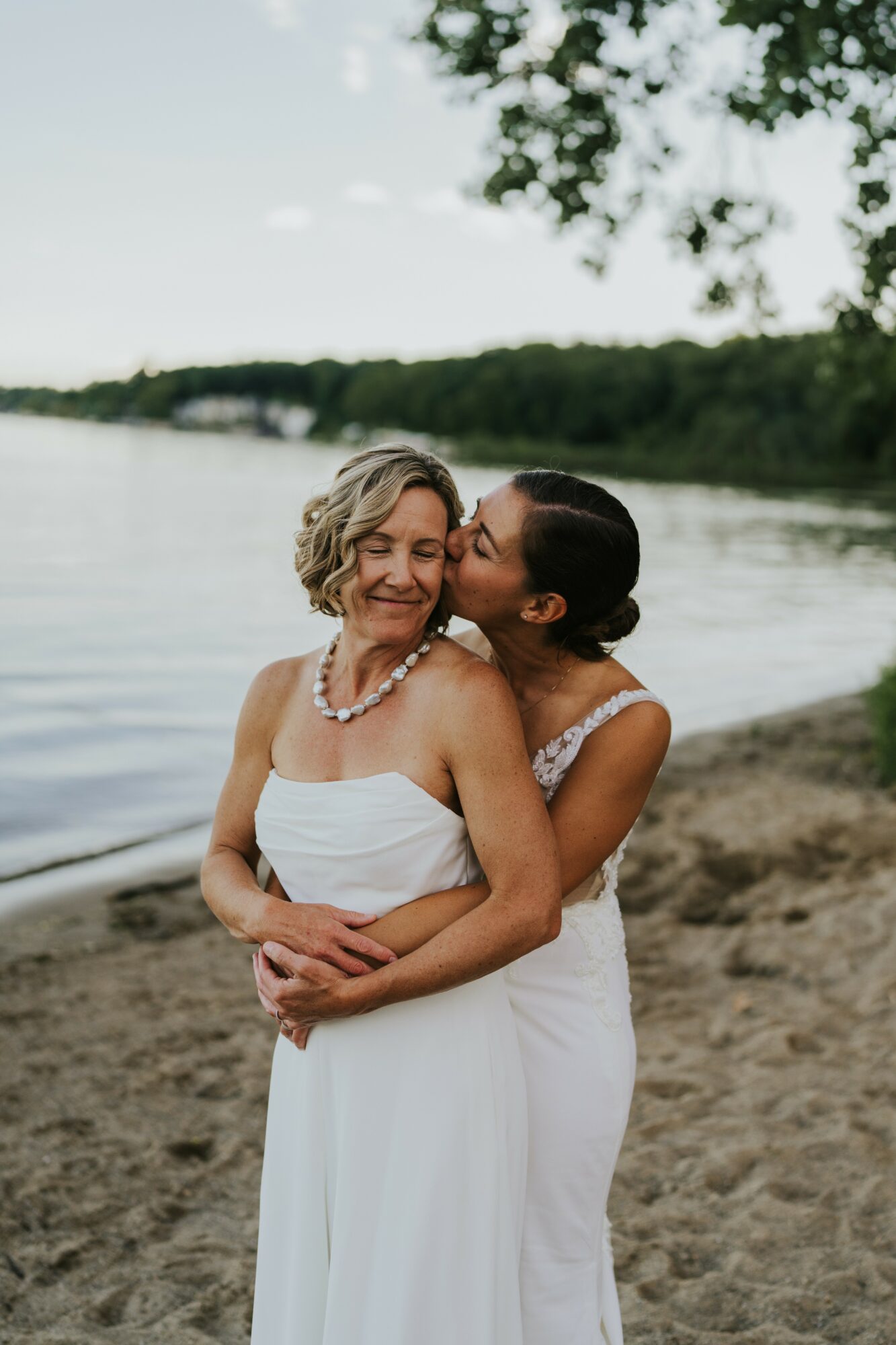 Two women in white dresses embrace on a beach, one kissing the other's cheek, with water and trees in background.