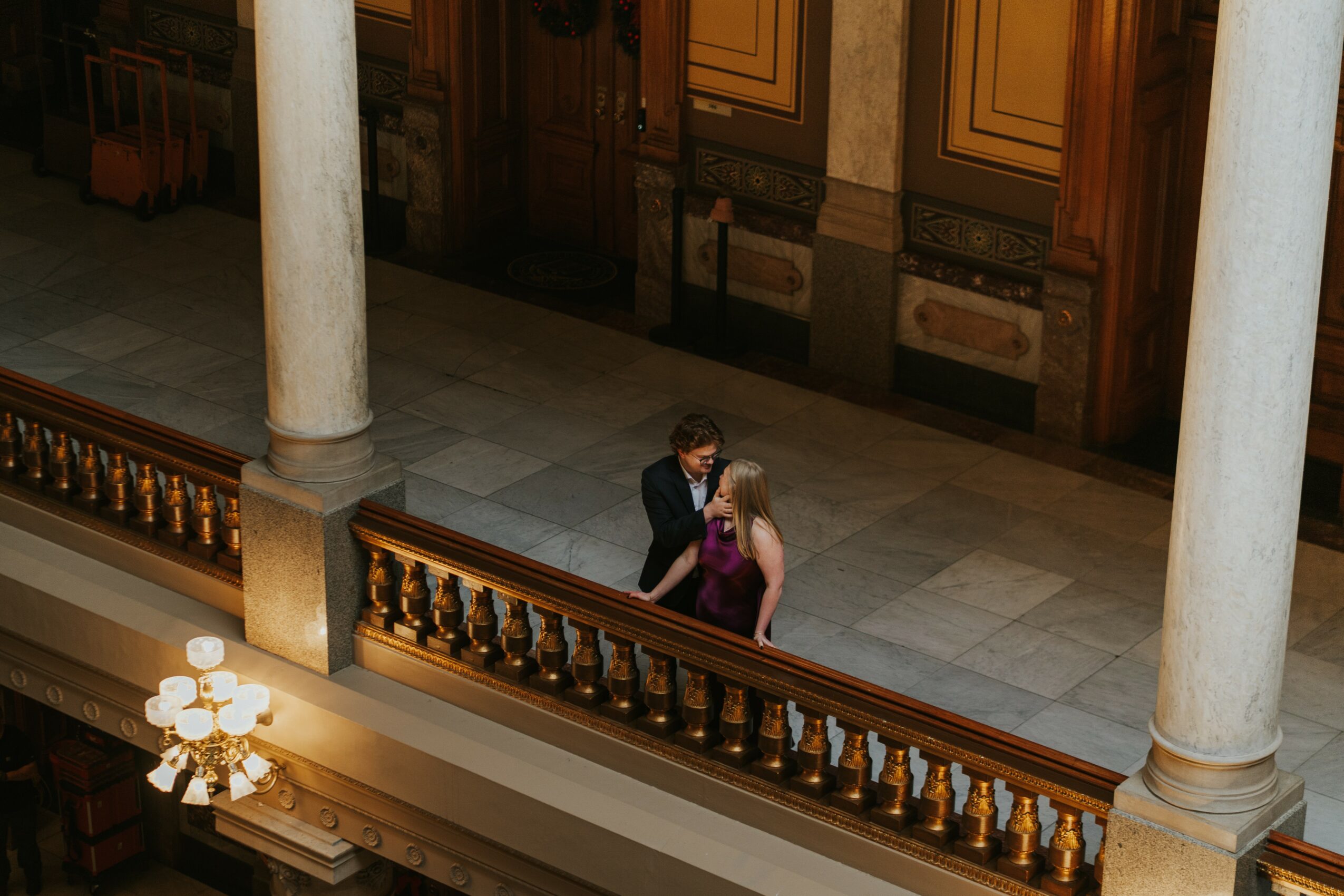 Two people stand on a balcony with ornate wooden railing, in a grand interior with columns and decorative wall panels.