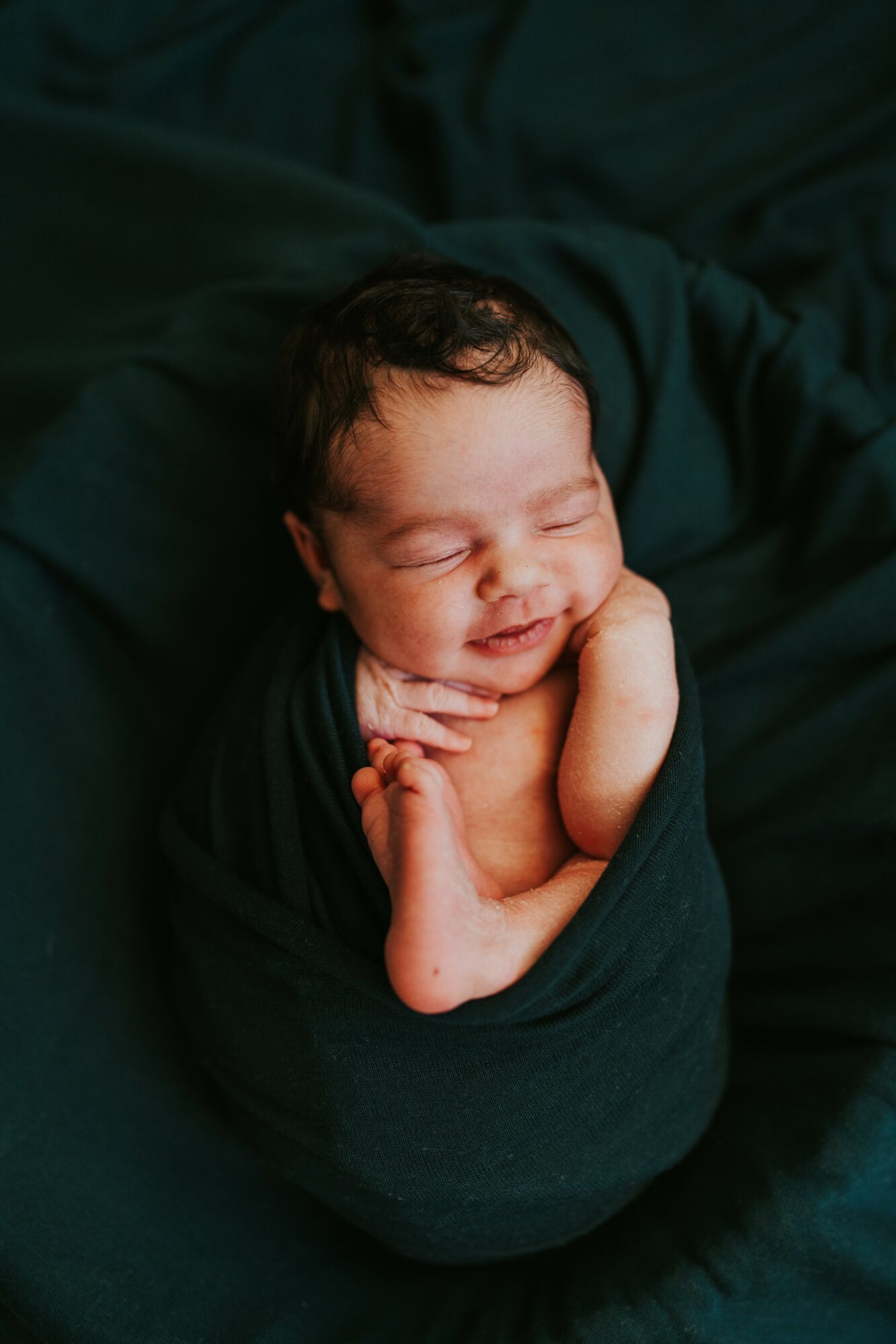 Newborn baby wrapped in dark fabric, sleeping peacefully with hand near face, on dark background.
