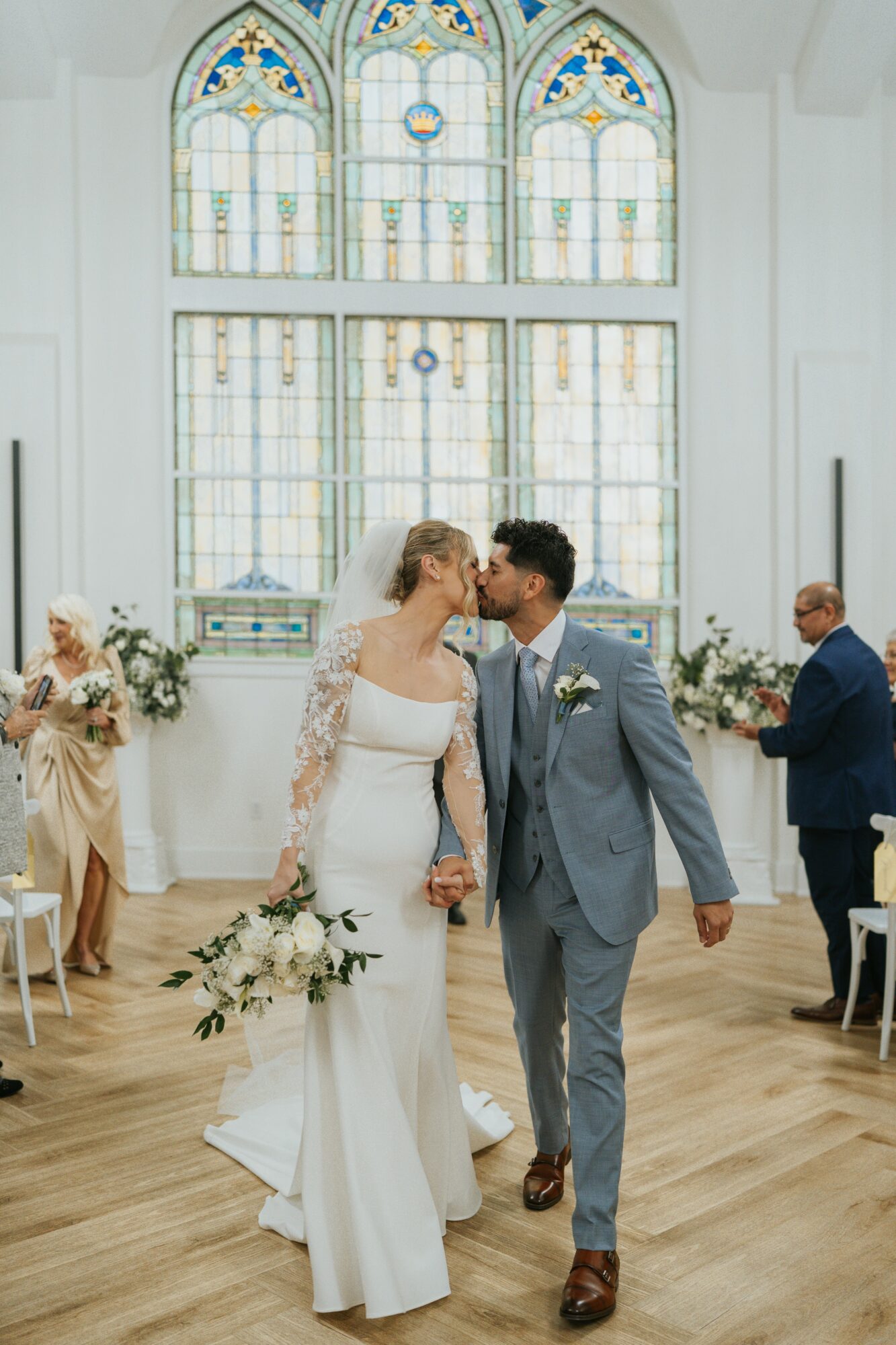 Bride and groom kiss in a church with stained glass windows, holding hands, surrounded by guests and floral arrangements.
