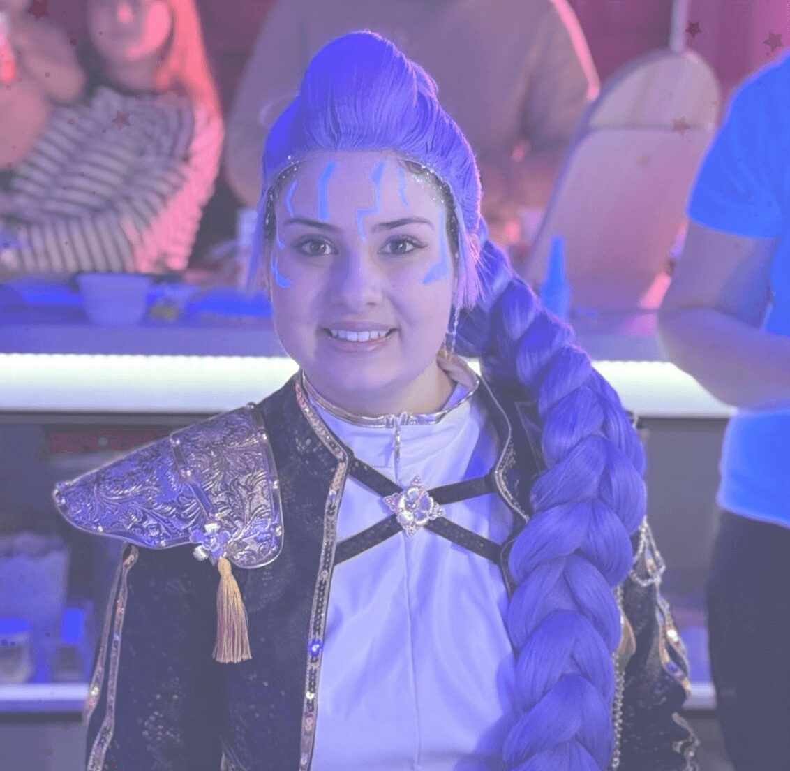 Young woman with long purple braided hair, wearing a costume with metallic shoulder pads, smiling at the camera.