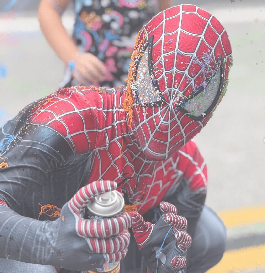 Person in a Spider-Man costume with a damaged mask, holding a spray can, outdoors, with a blurred background.