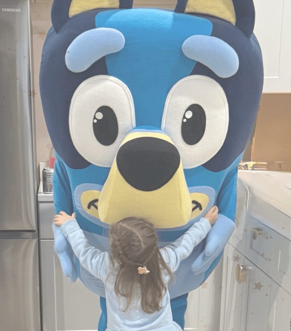 Child with long hair and a pink hair clip reaches out to a large cartoon character costume with big eyes and a yellow nose.