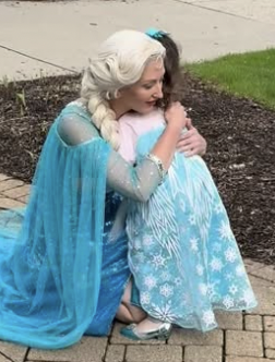 Woman dressed as Elsa from Frozen hugging a child in a blue dress outdoors, on a brick path with grass and soil nearby.