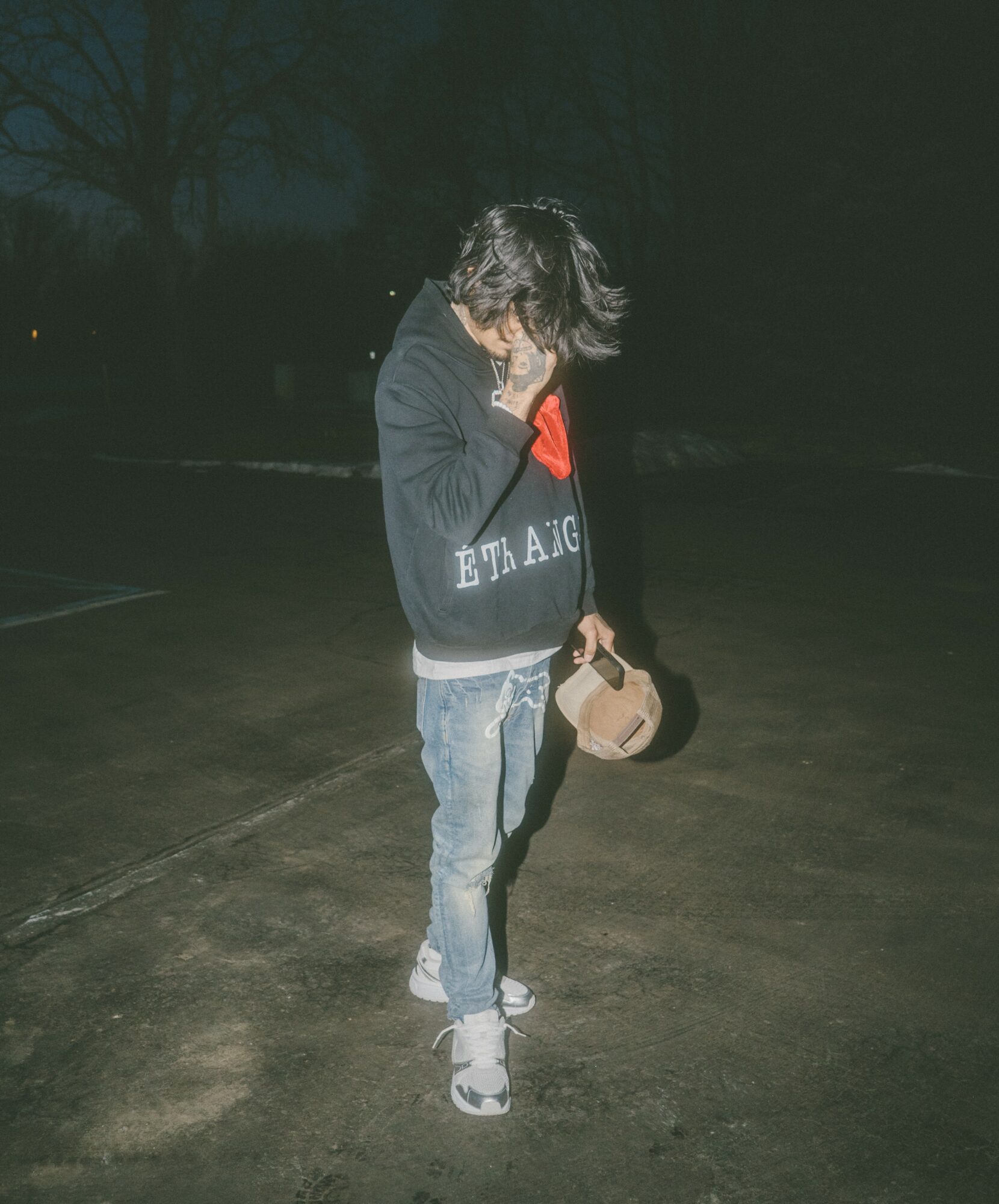 Person standing outdoors at night, wearing a hoodie, jeans, and sneakers, holding a hat, with hair covering face.