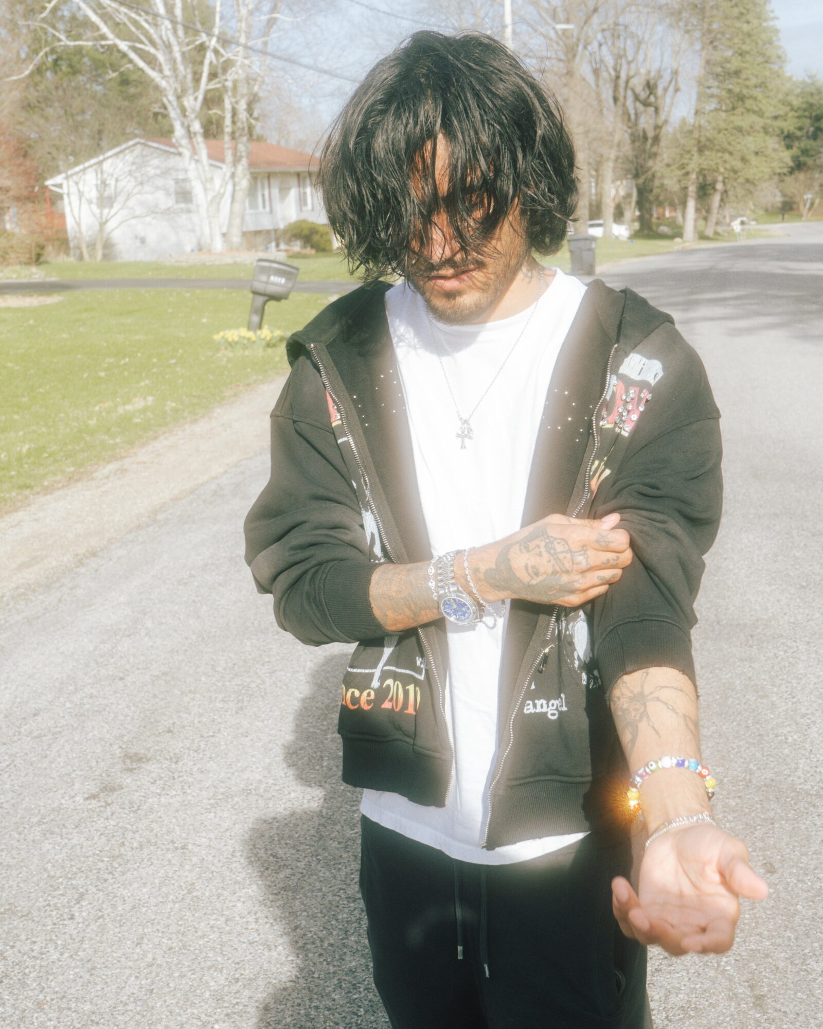 Young man with dark curly hair, wearing sunglasses, a black hoodie, and white shirt, standing outdoors on a sunny day.