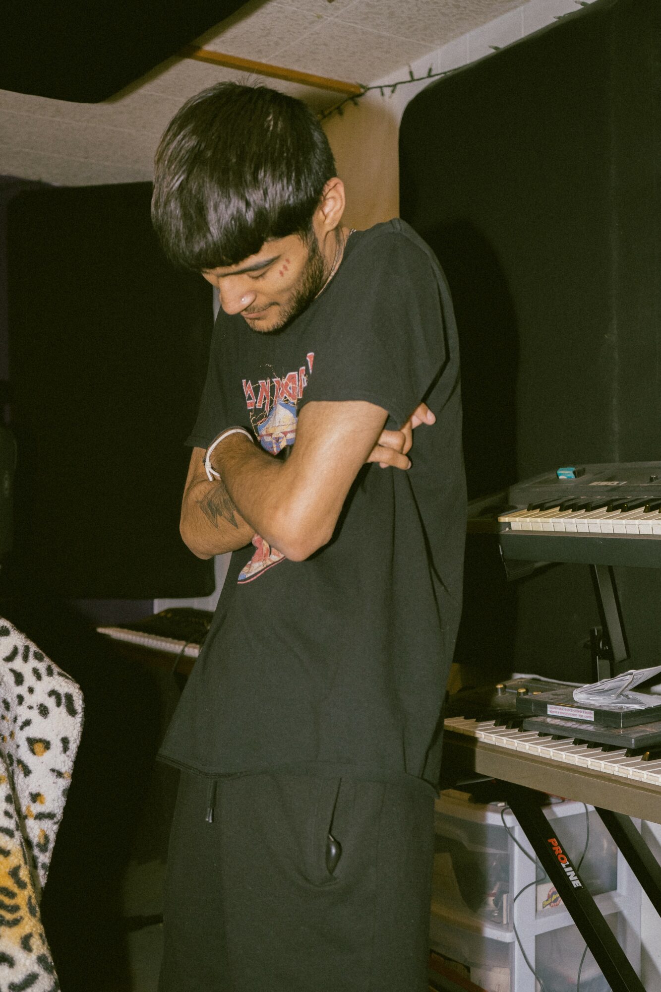 Young person with dark hair and black clothing stands with arms crossed in a music studio, surrounded by keyboards and equipment.
