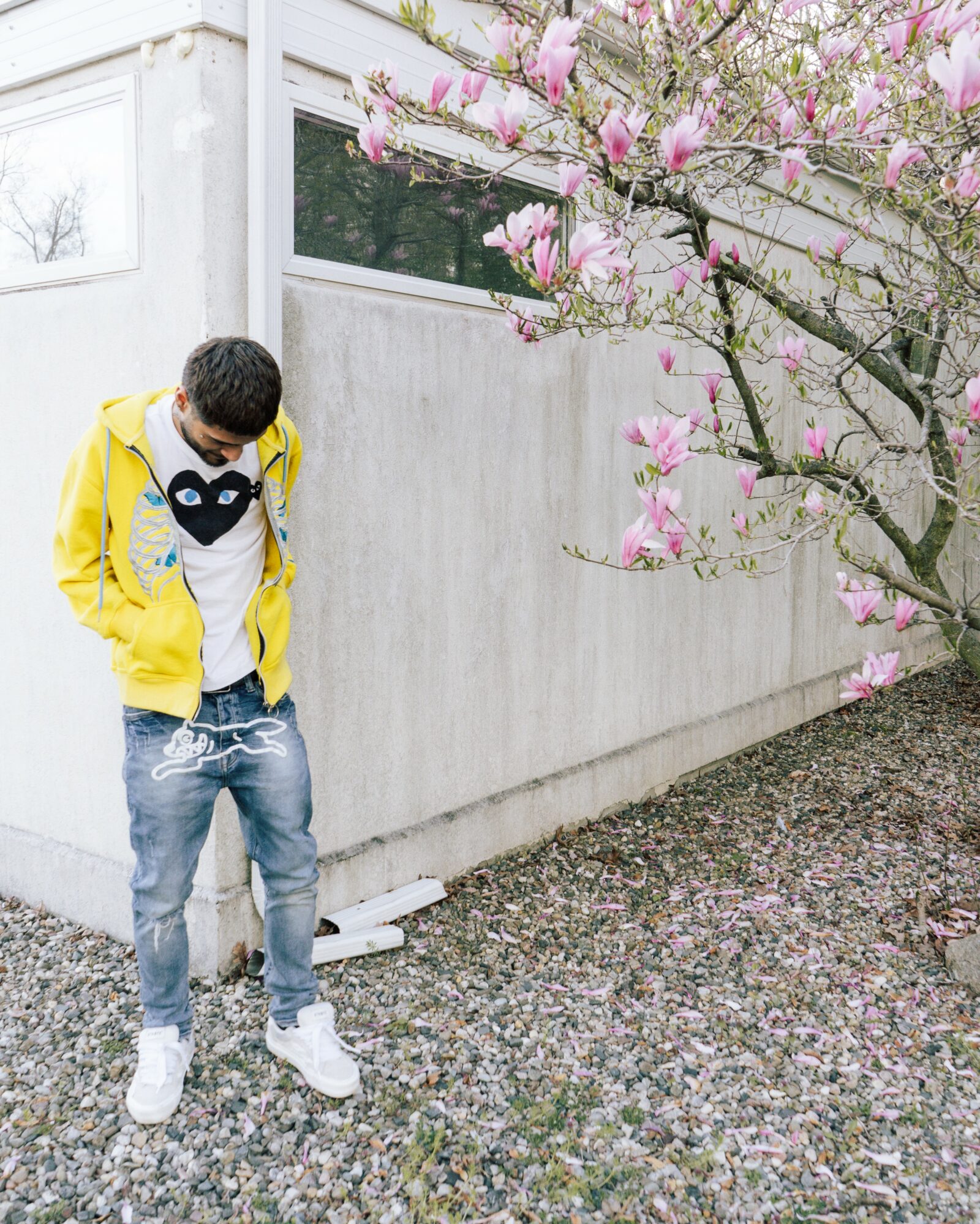 Young person in yellow jacket and jeans stands outdoors near a flowering tree with pink blossoms, against a concrete wall.
