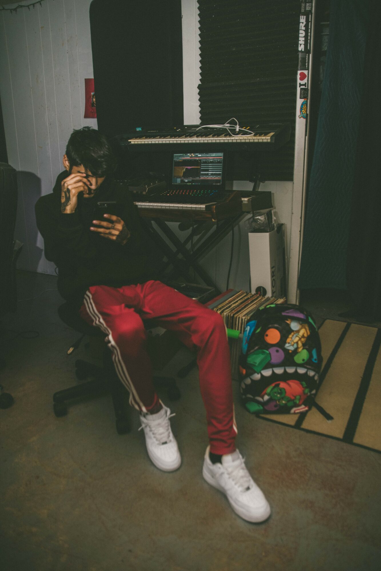 Person sitting in a music studio, wearing red pants and white sneakers, with a backpack nearby.