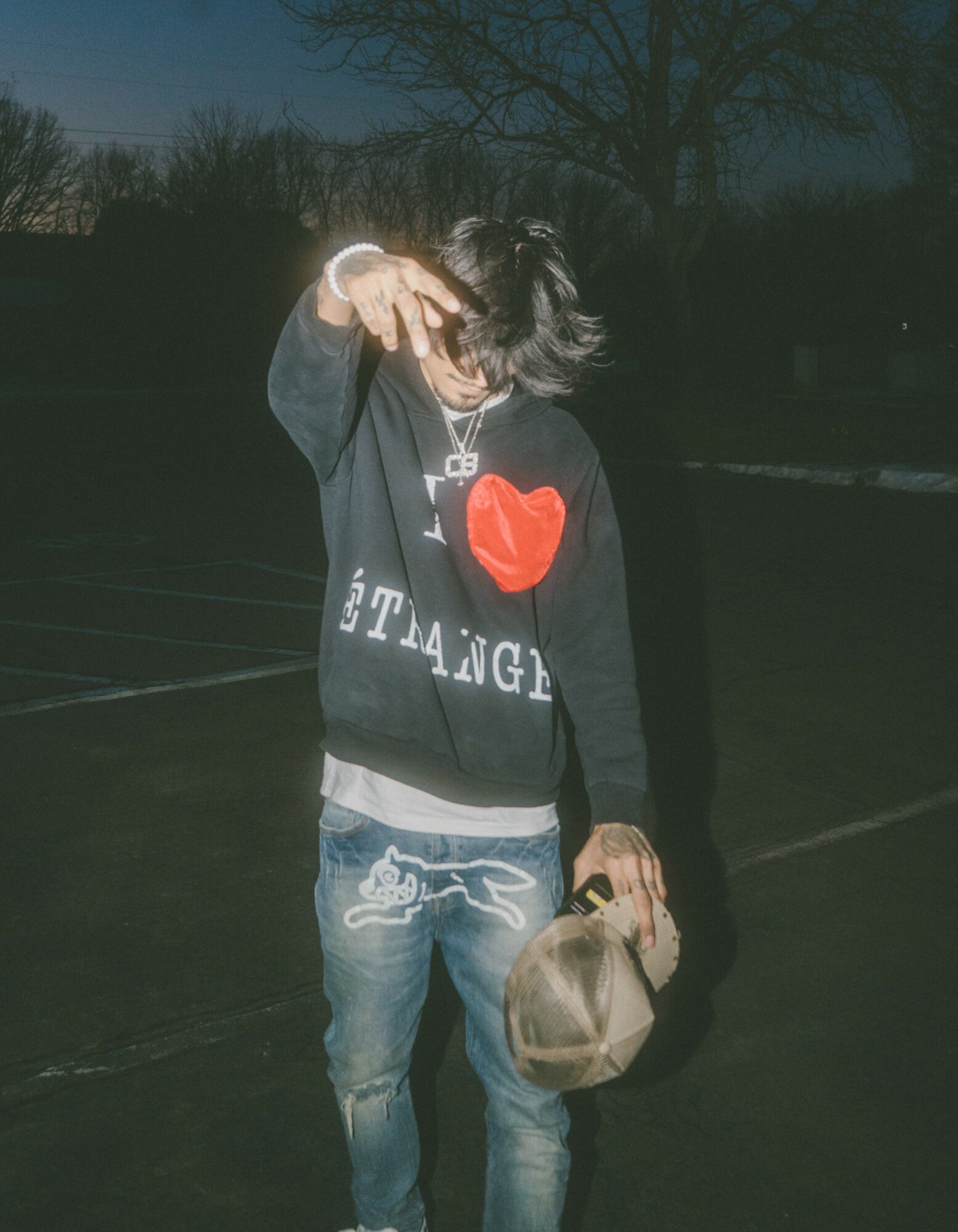 Person with dark hair wearing a black hoodie with a red heart and text, holding a bag, standing outdoors at night.