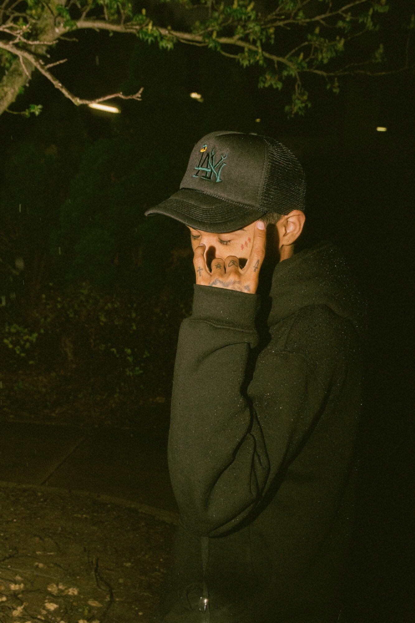 Person wearing a black cap and hoodie, covering their face with their hand, outdoors at night with trees in background.