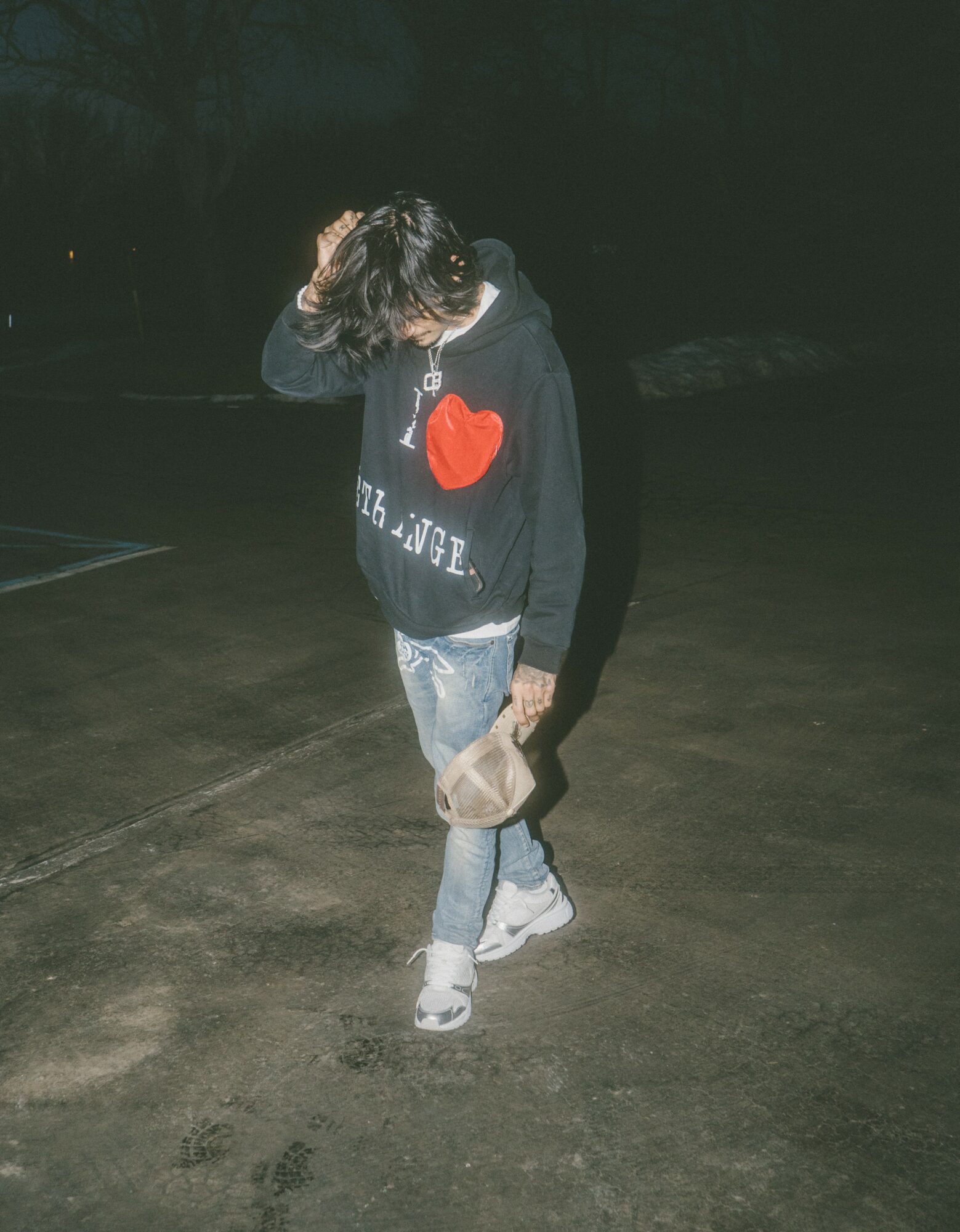 Person walking outdoors at night, wearing a black hoodie with a red heart, jeans, and white shoes, holding a beige bag.