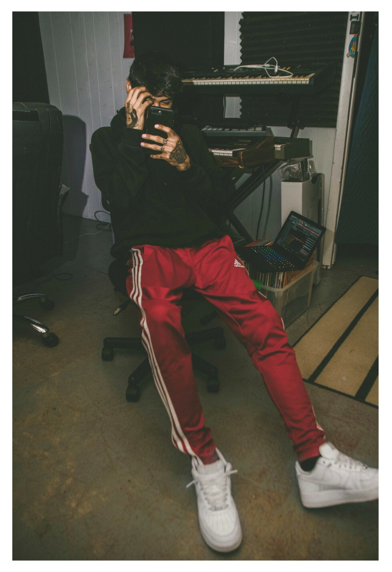 Person taking a mirror selfie in a music studio, wearing a black hoodie, red pants, and white sneakers.