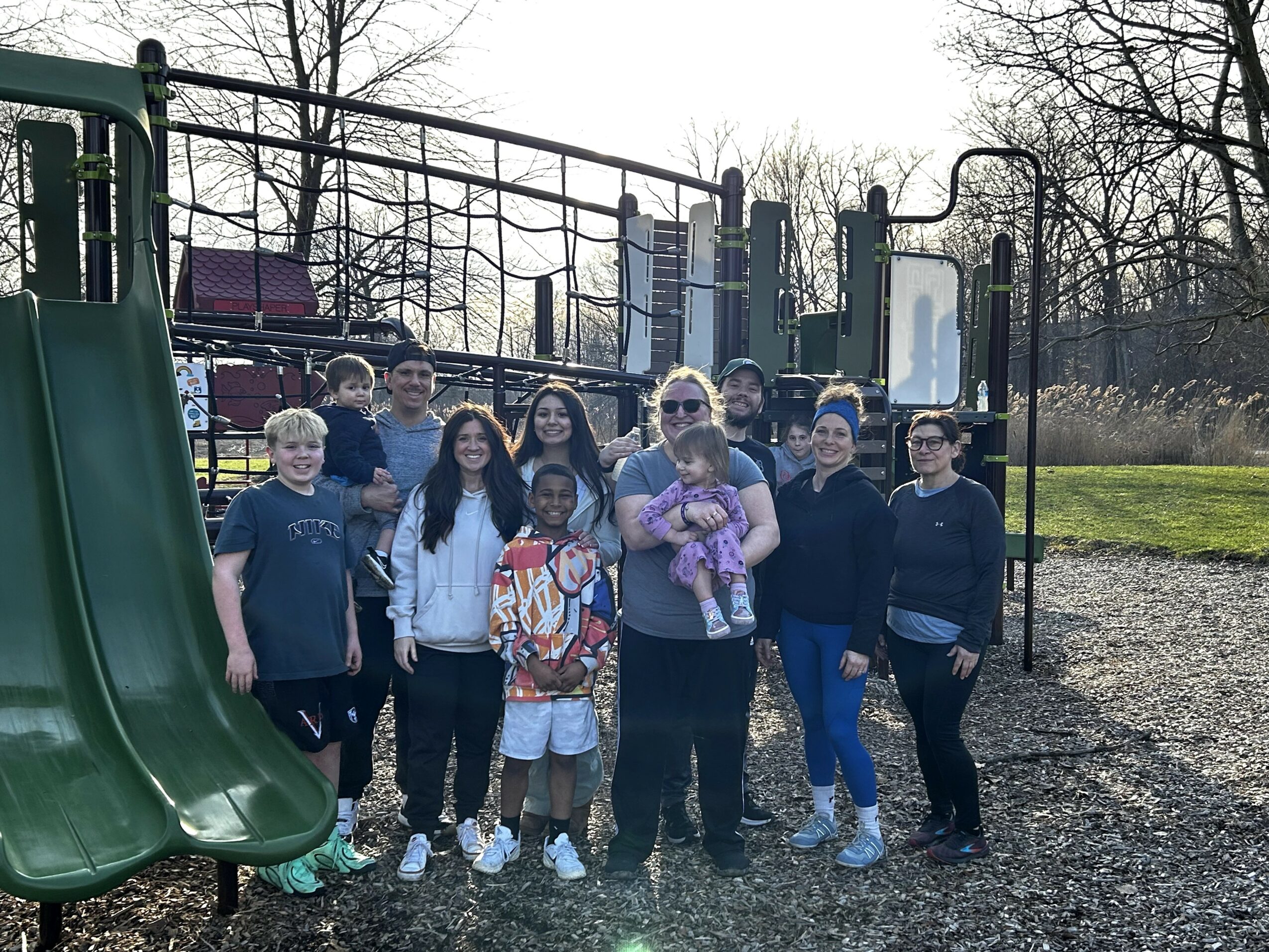 Group of children and adults standing on playground with slides and trees in background.