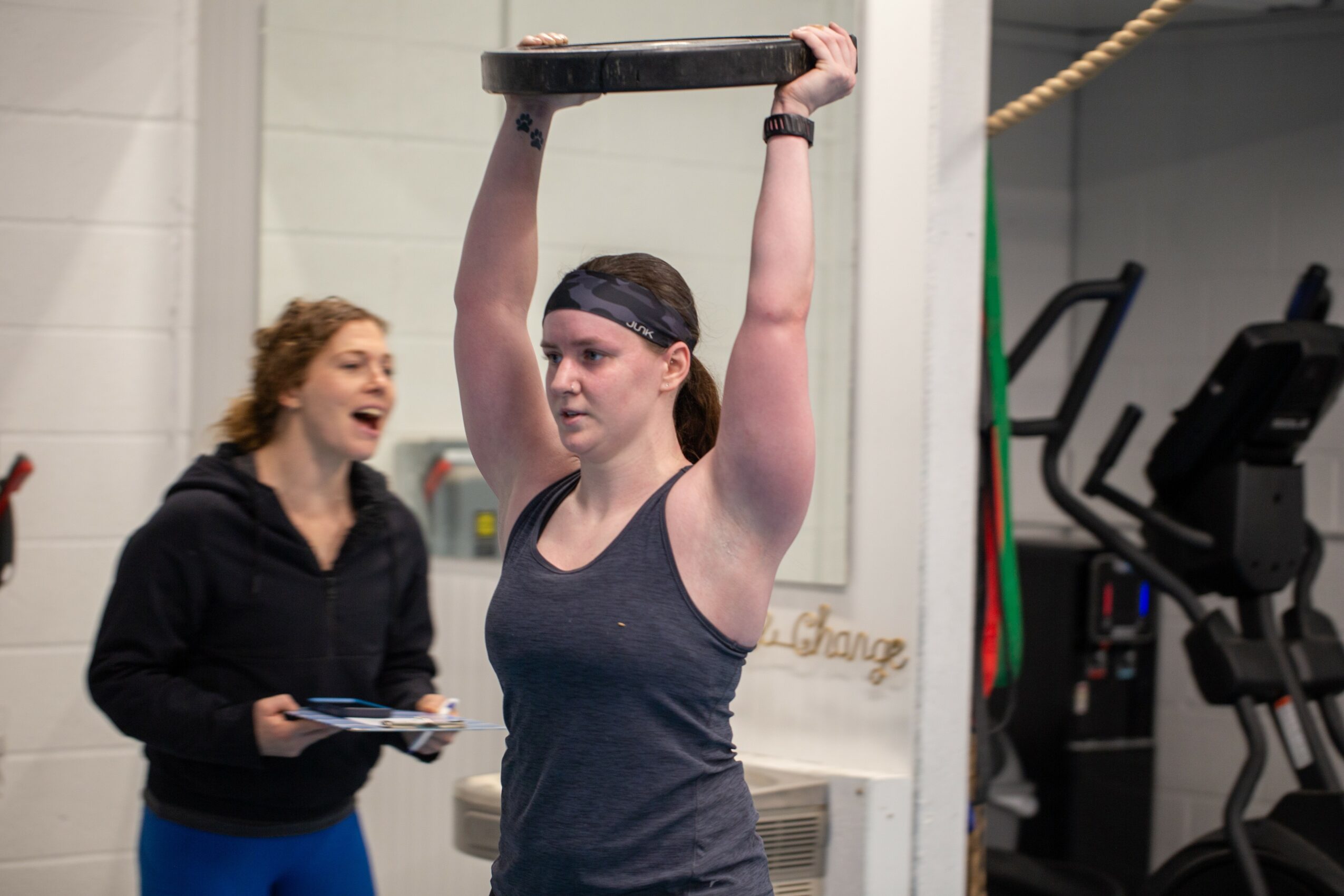 Woman lifting a weight plate overhead during workout, smiling woman in background, gym setting, fitness activity