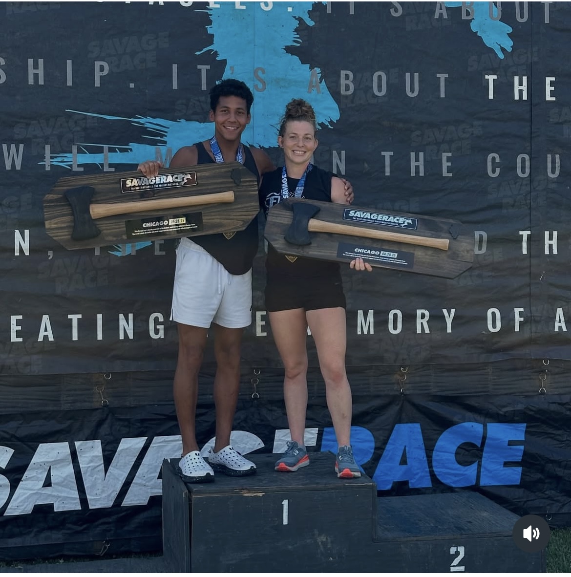 Two young people standing on a winners' podium holding large wooden plaques, smiling, with a world map background.
