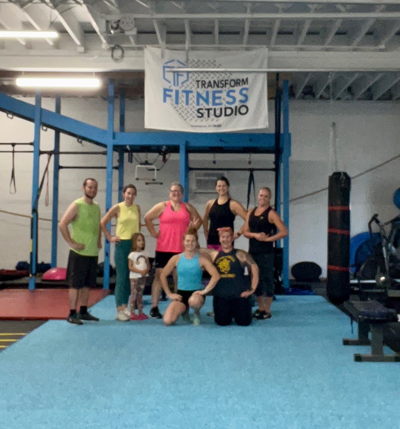 Group of seven people in gym, some standing, some kneeling, in front of fitness equipment and a sign that reads 'Transform Fitness Studio'.