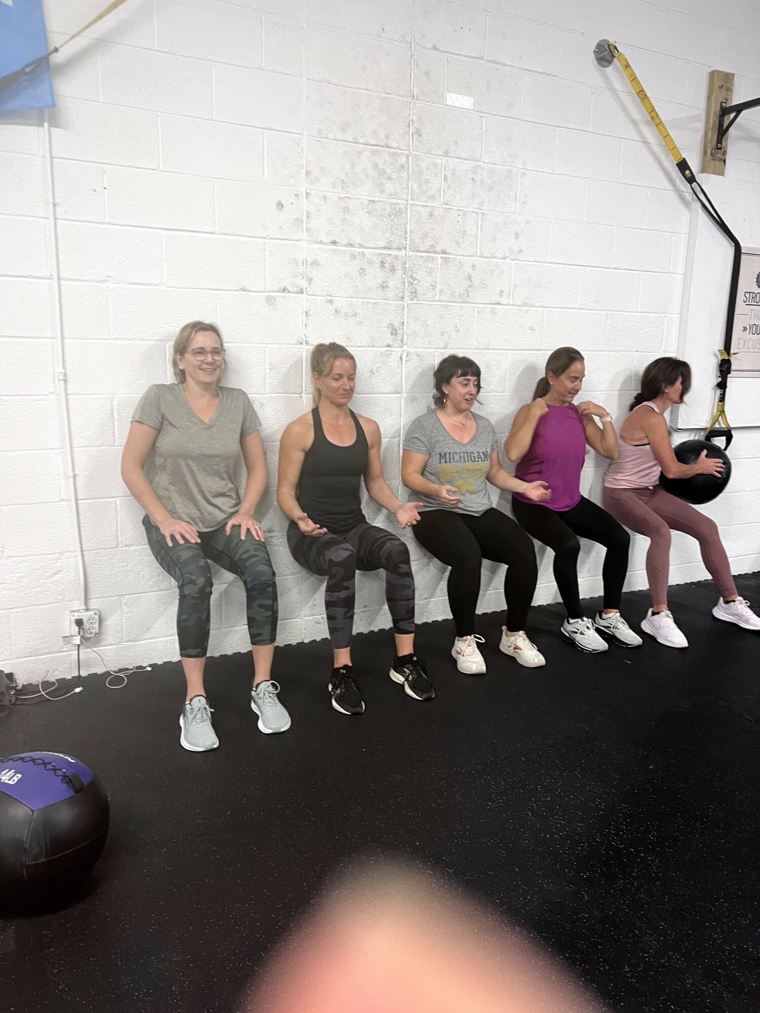 Five women sit on a wall in a gym, with a medicine ball on the floor and gym equipment on the wall behind them.
