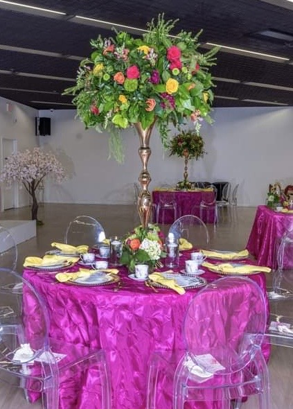 Round table with a bright pink tablecloth, set with yellow napkins, white plates, and glassware, decorated with a tall floral centerpiece.