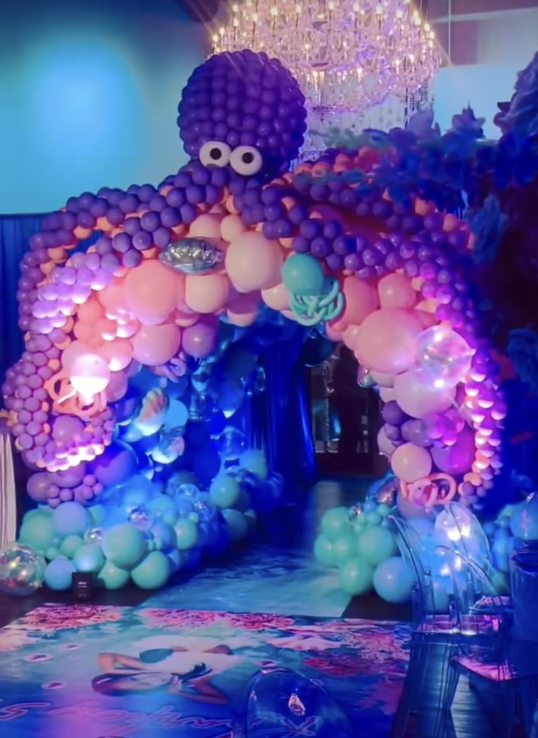 Colorful balloon arch with purple, pink, and blue balloons, decorated with a large purple balloon cluster and a chandelier above.