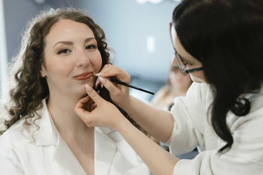 Makeup artist applies lipstick to a woman with curly hair in a white shirt, in a well-lit room.