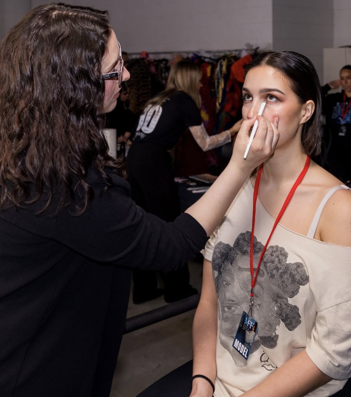 Makeup artist applies makeup to a young woman with a lanyard, in a busy backstage area.