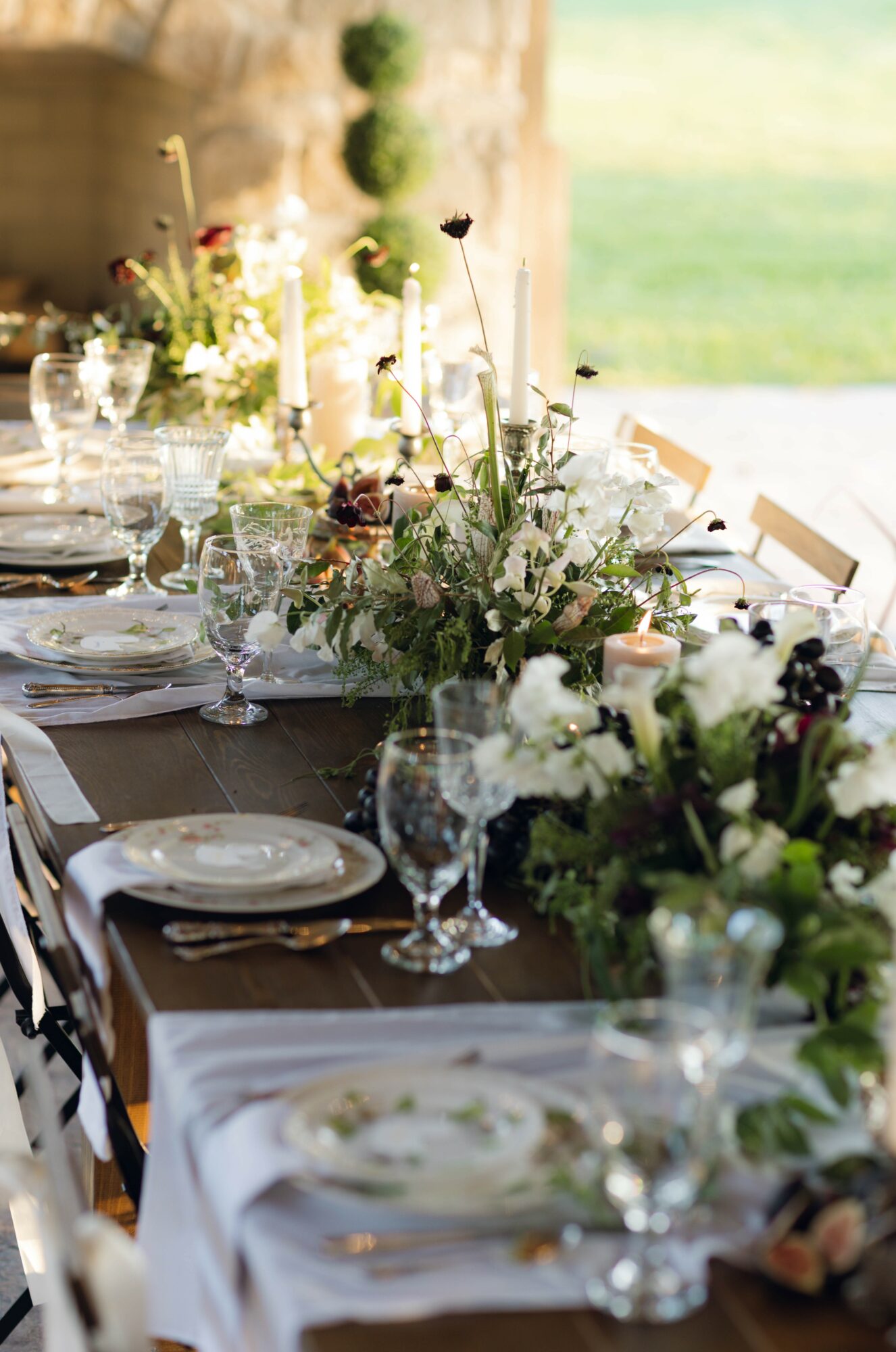 Elegant table setting with white flowers, candles, glassware, and plates in a bright outdoor setting.