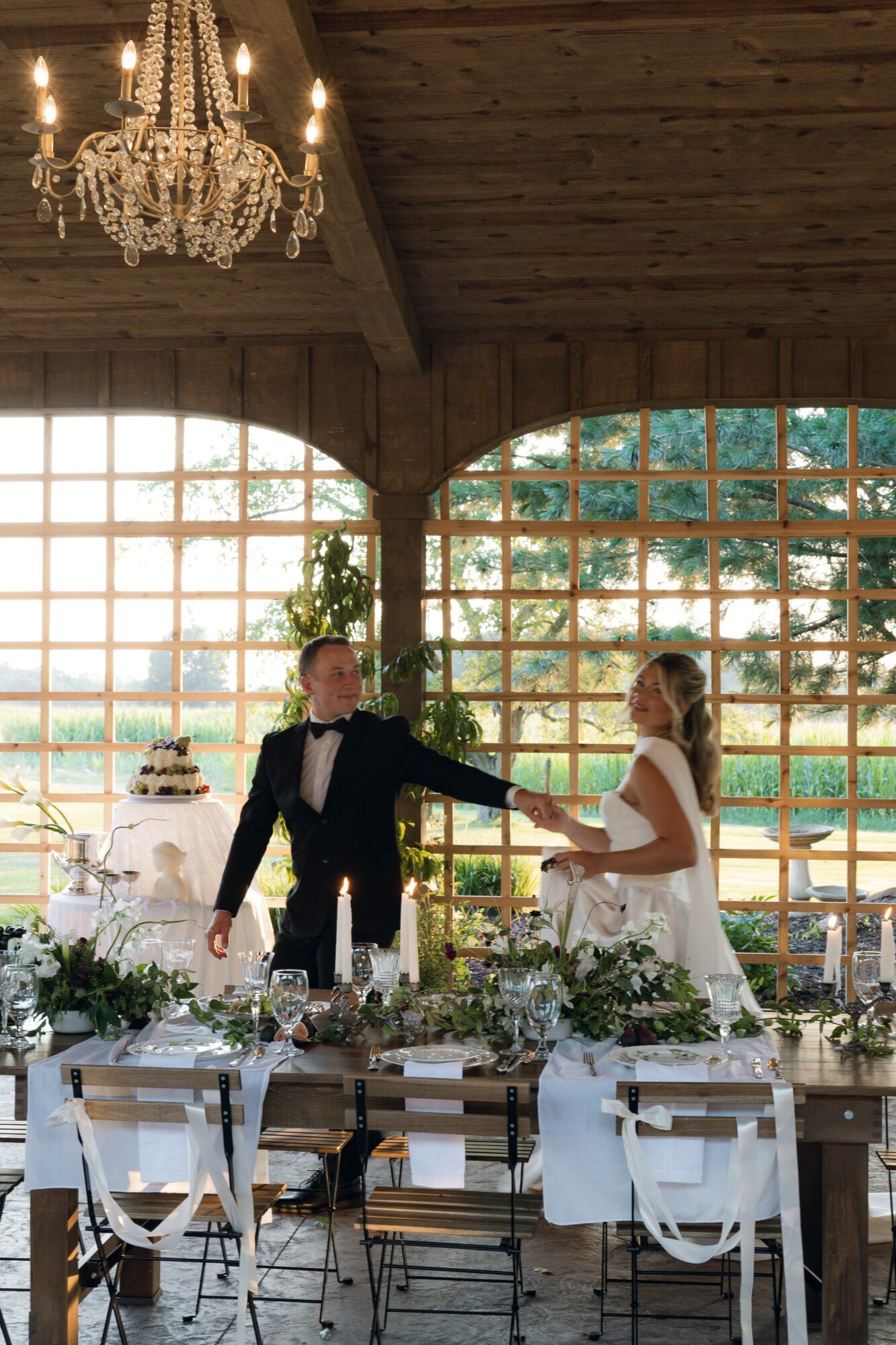 A bride and groom dance indoors with large windows and a chandelier, decorated with flowers and a wedding cake nearby.