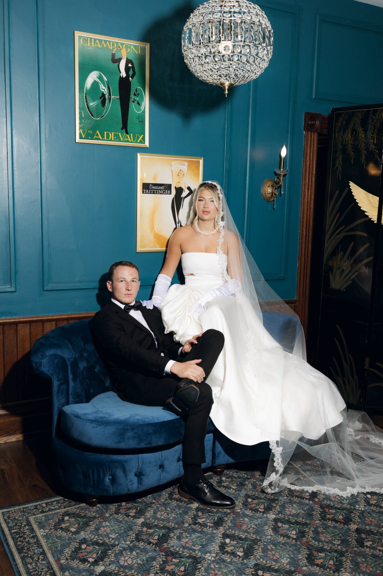 Bride in white gown and veil stands beside groom in black tuxedo on blue velvet sofa in elegant room with chandelier and artwork.