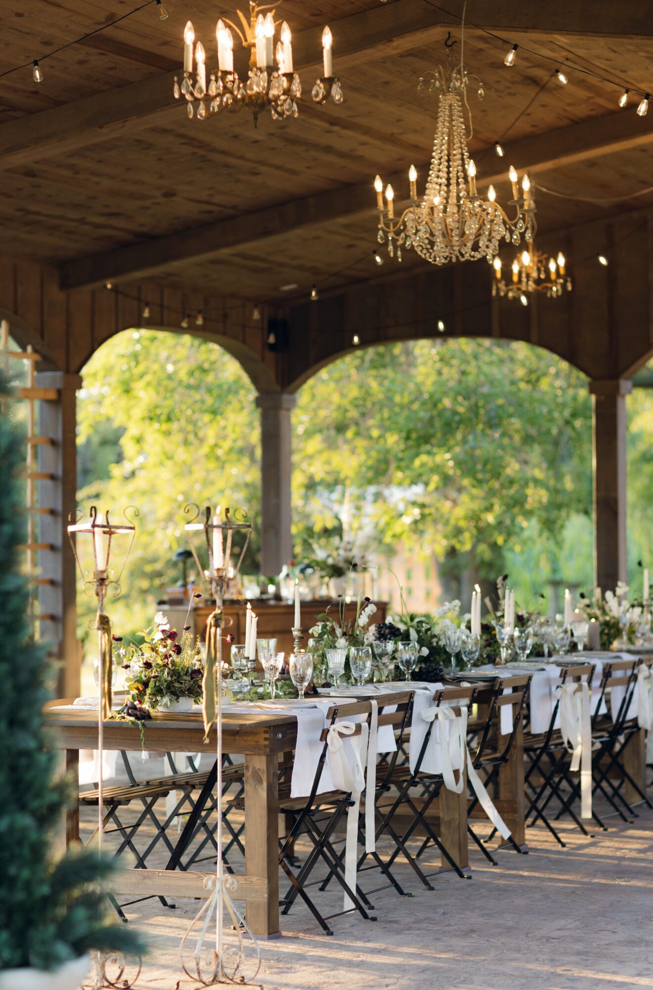 Long outdoor dining table with chairs, decorated with candles and greenery, under a wooden pavilion with chandeliers, surrounded by trees