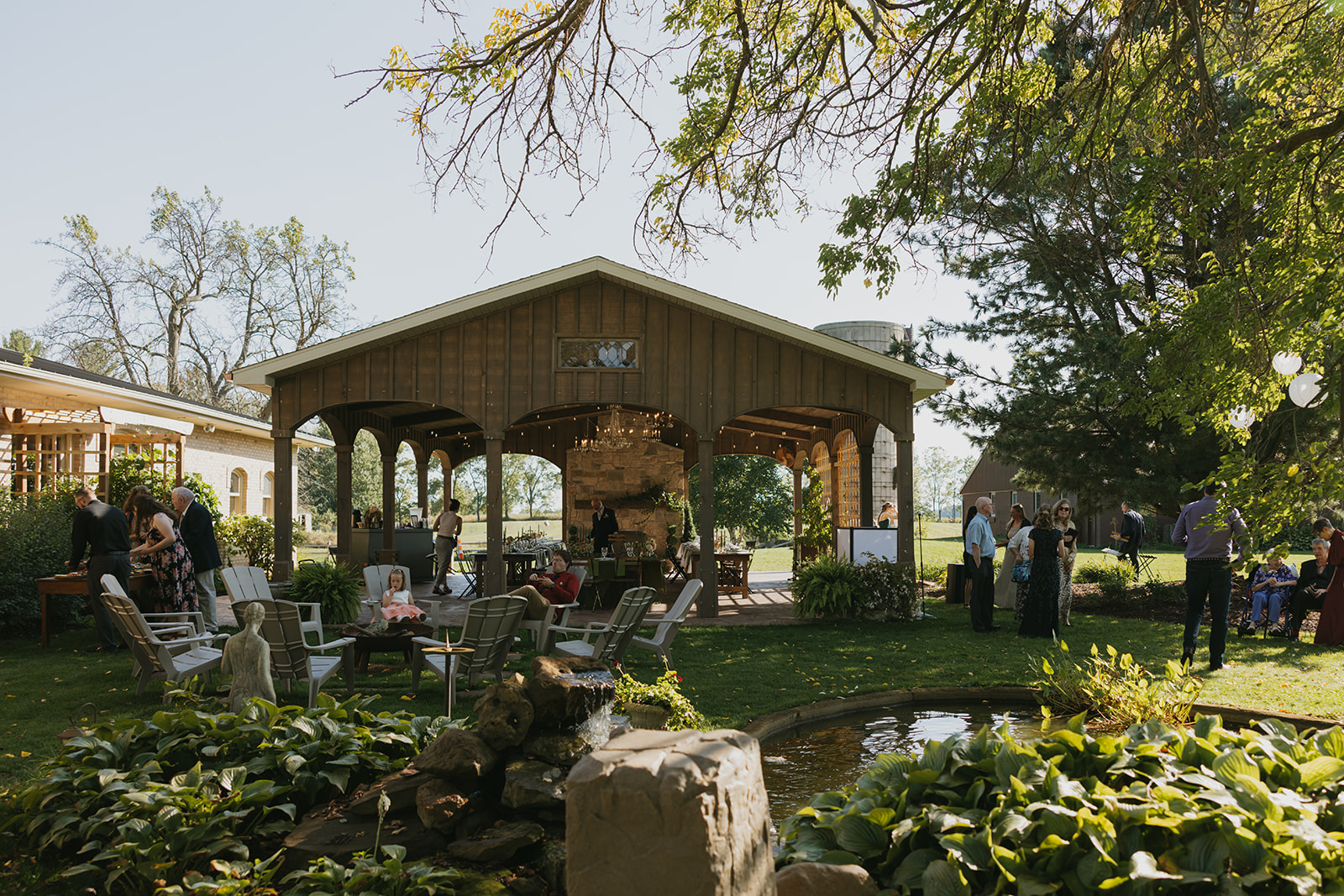 People gather around a pavilion with a brick fireplace, surrounded by trees and outdoor furniture in a garden setting.