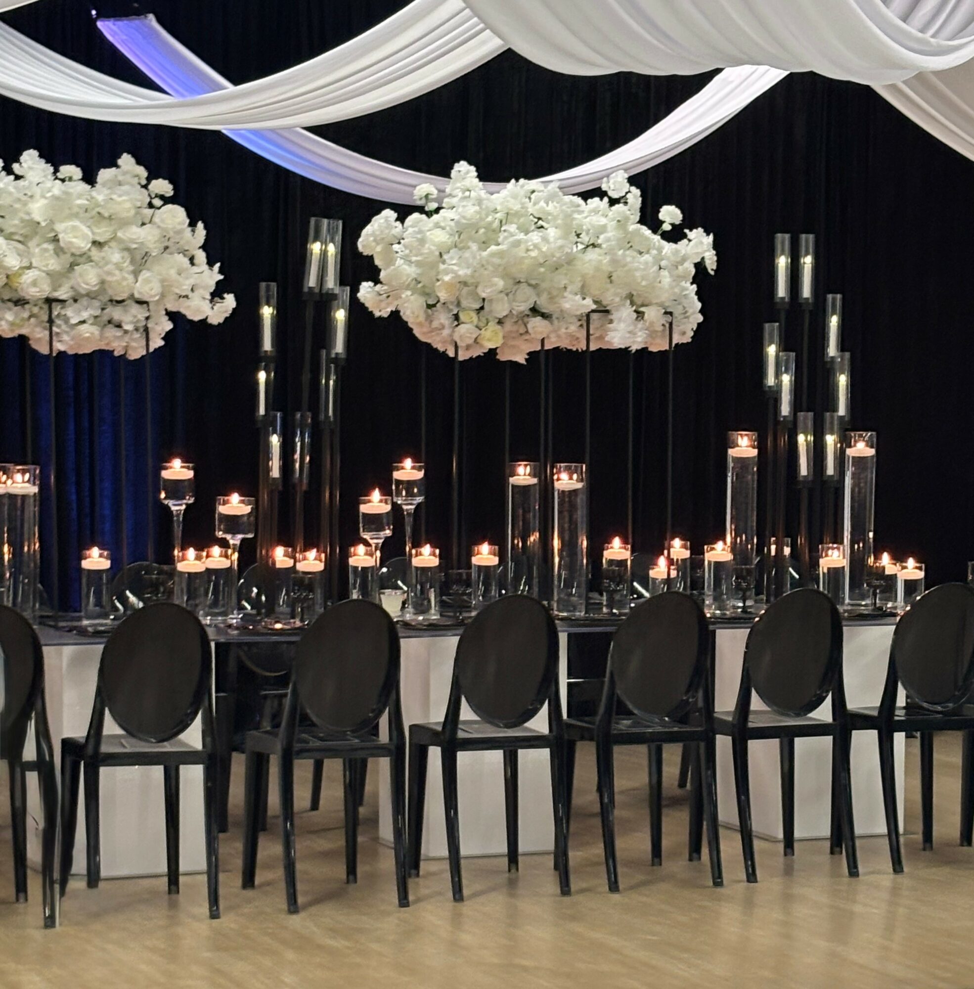 Elegant event setup with black chairs, floral arrangements, and tall candles on a table, black and white drapery overhead.