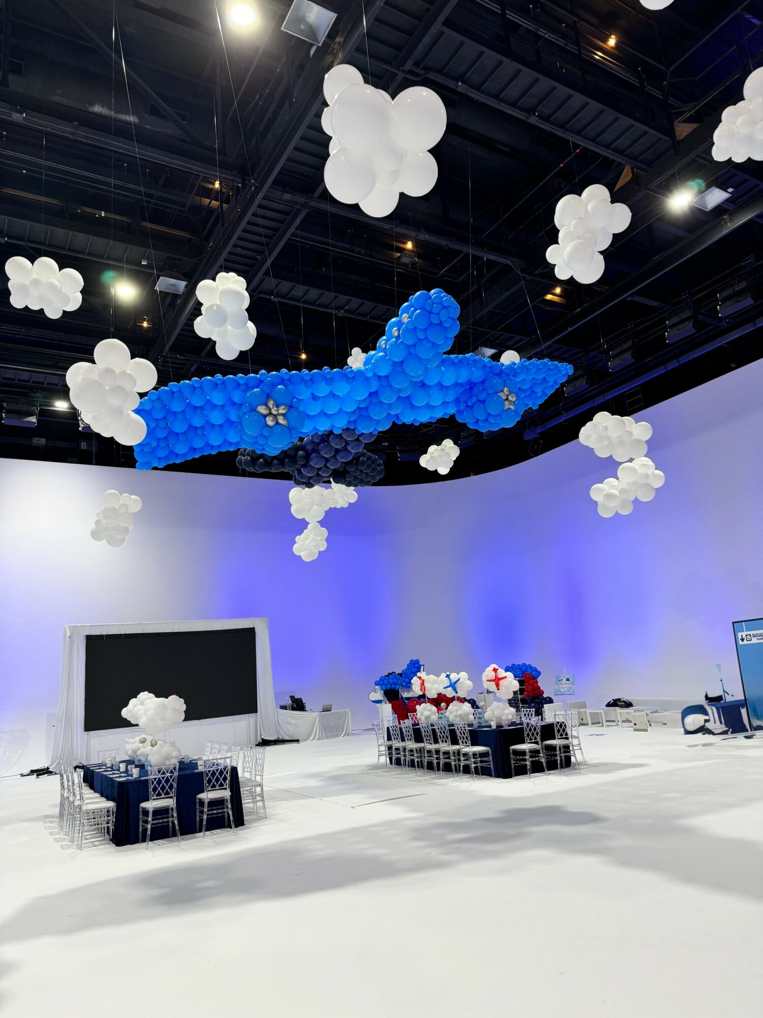 Indoor event space with cloud and airplane balloon decorations, tables with chairs, and a large screen, illuminated with blue lighting.