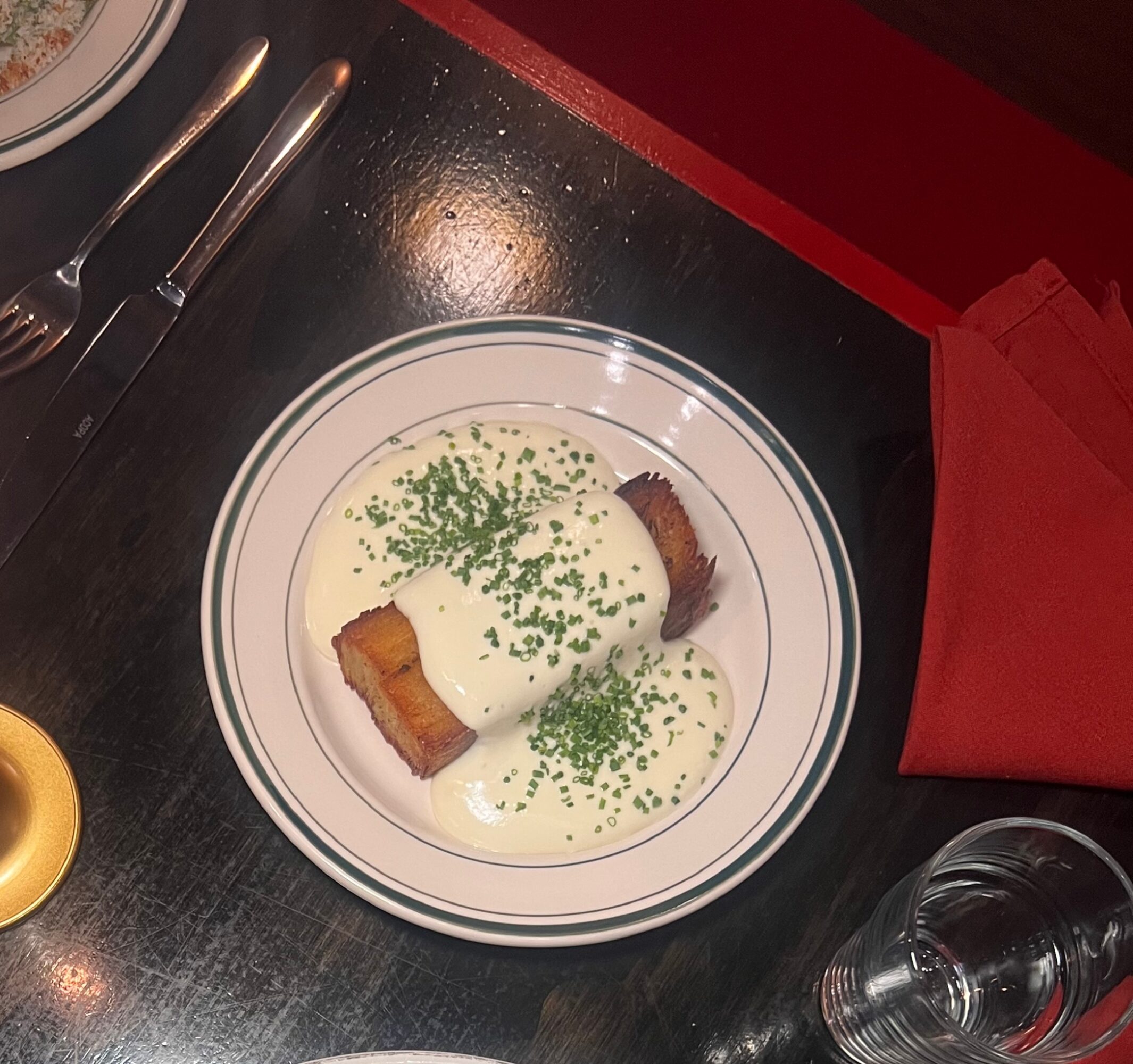 Plate with two pieces of meat topped with white sauce and garnished with chopped herbs, on a dark table with utensils and a glass nearby.