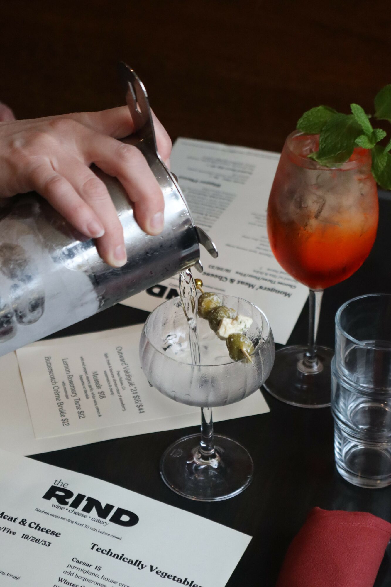 Person pouring a drink into a cocktail glass with ice and garnishes, on a table with menus and a red napkin.