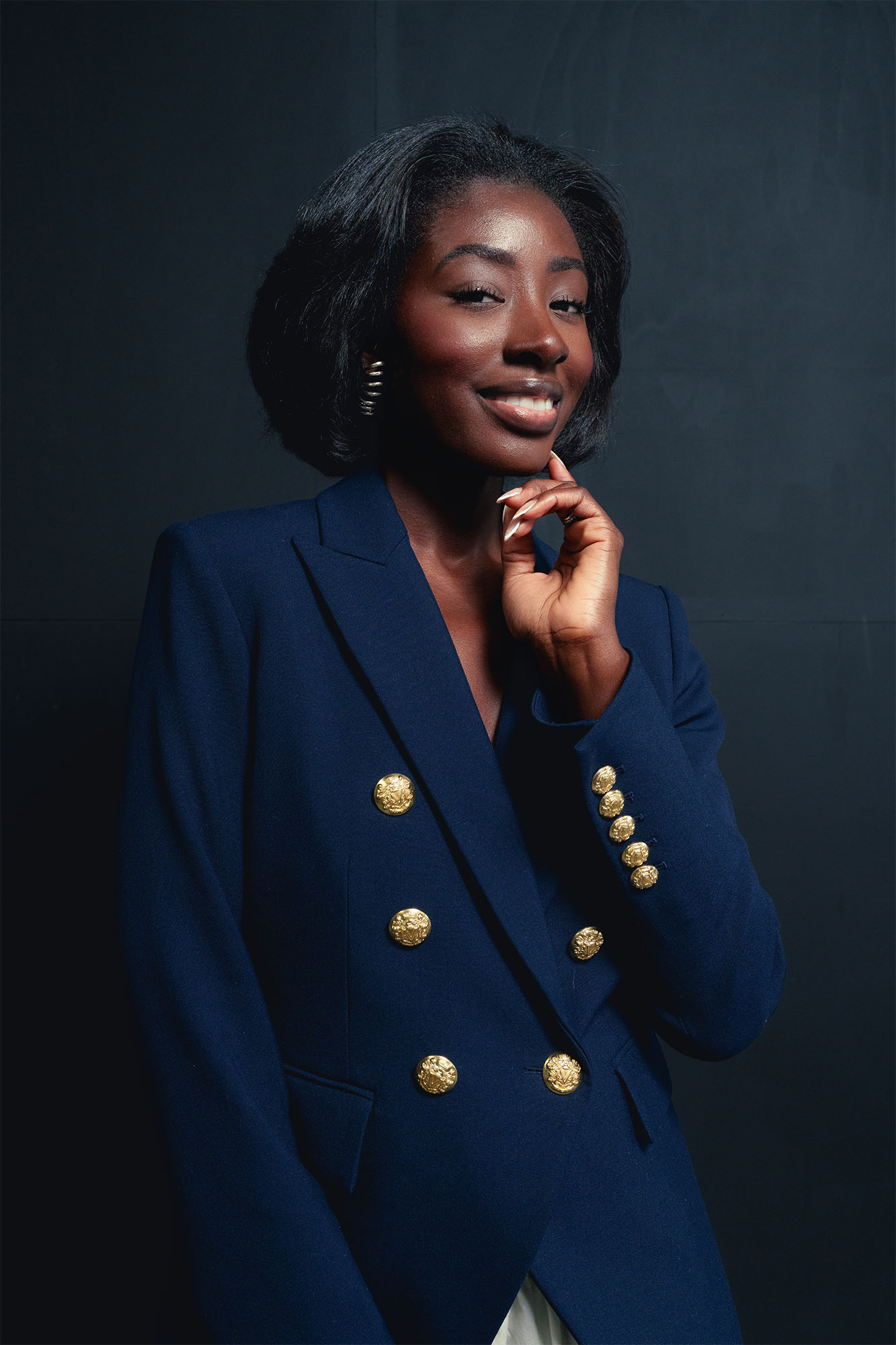 Young woman with short curly hair wearing a navy blazer with gold buttons, smiling and touching her chin, dark background.