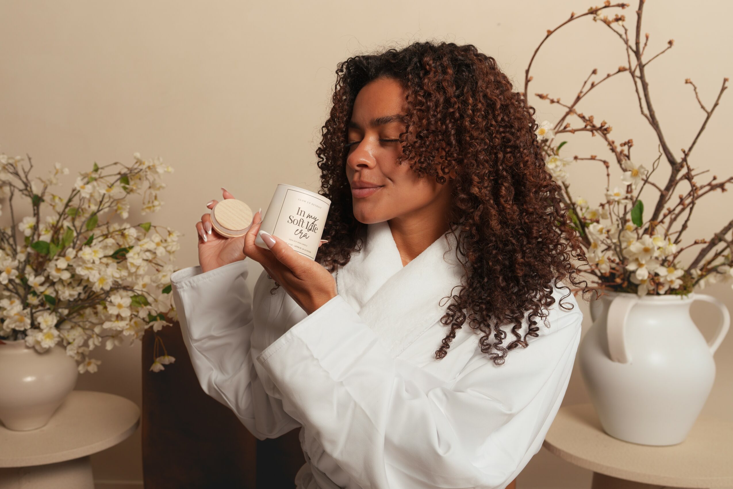 Woman with curly hair in a white robe holding a small jar, surrounded by vases with flowers and branches.