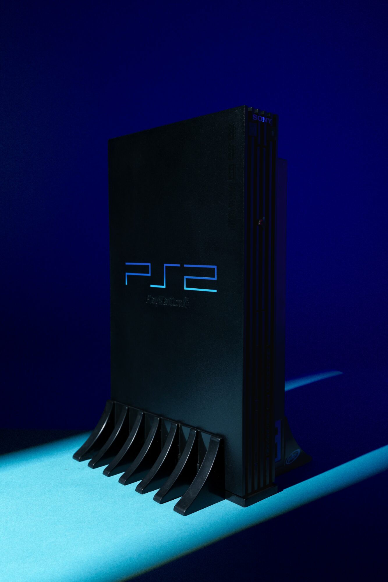 Vertical PlayStation 2 console with blue logo, standing on a surface with a dark background.