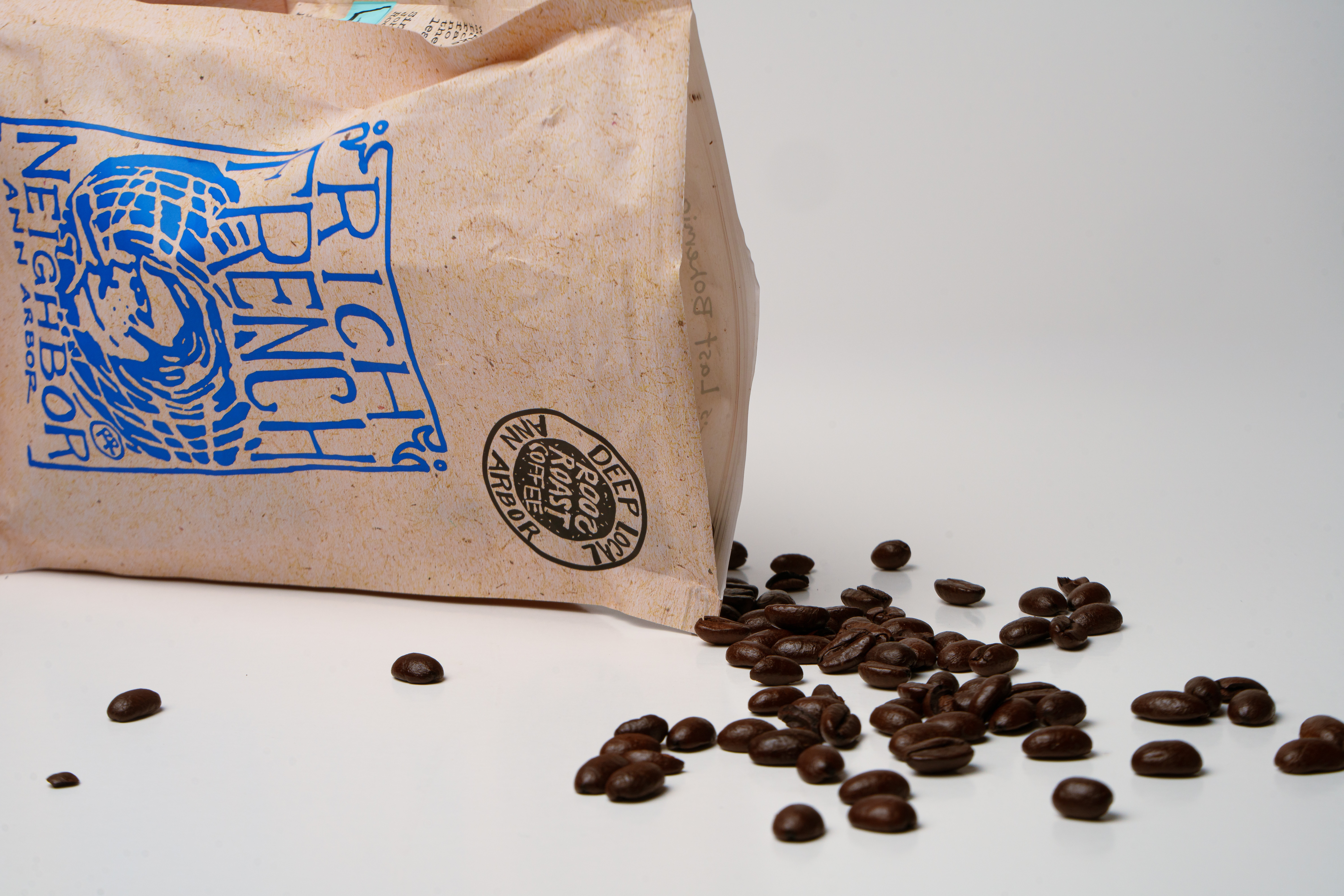 Coffee beans spilled from a paper bag onto a white surface, with the bag's blue logo visible.