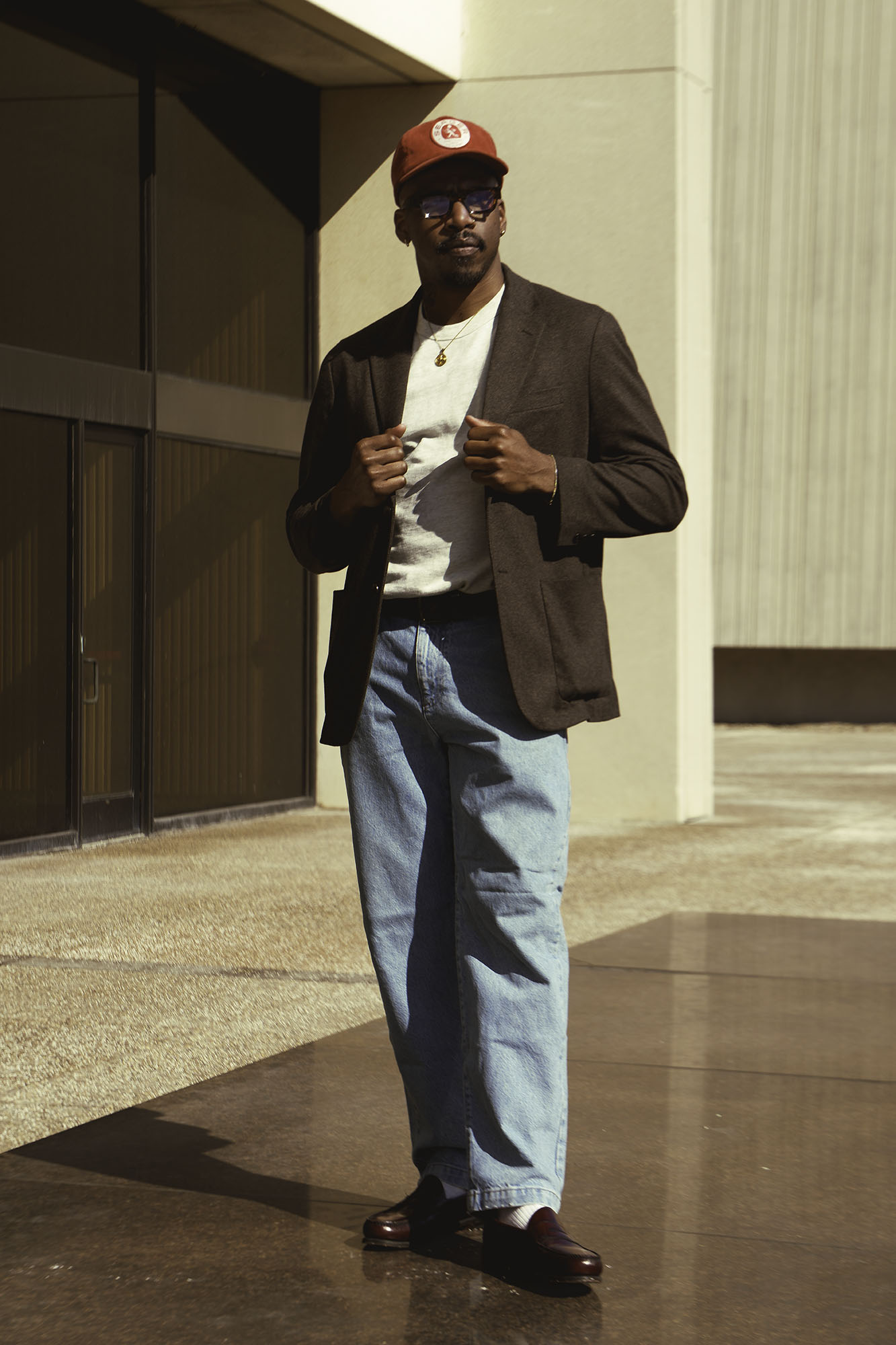 Man standing outdoors, wearing a red cap, sunglasses, brown blazer, white shirt, and blue jeans, holding a cigarette.