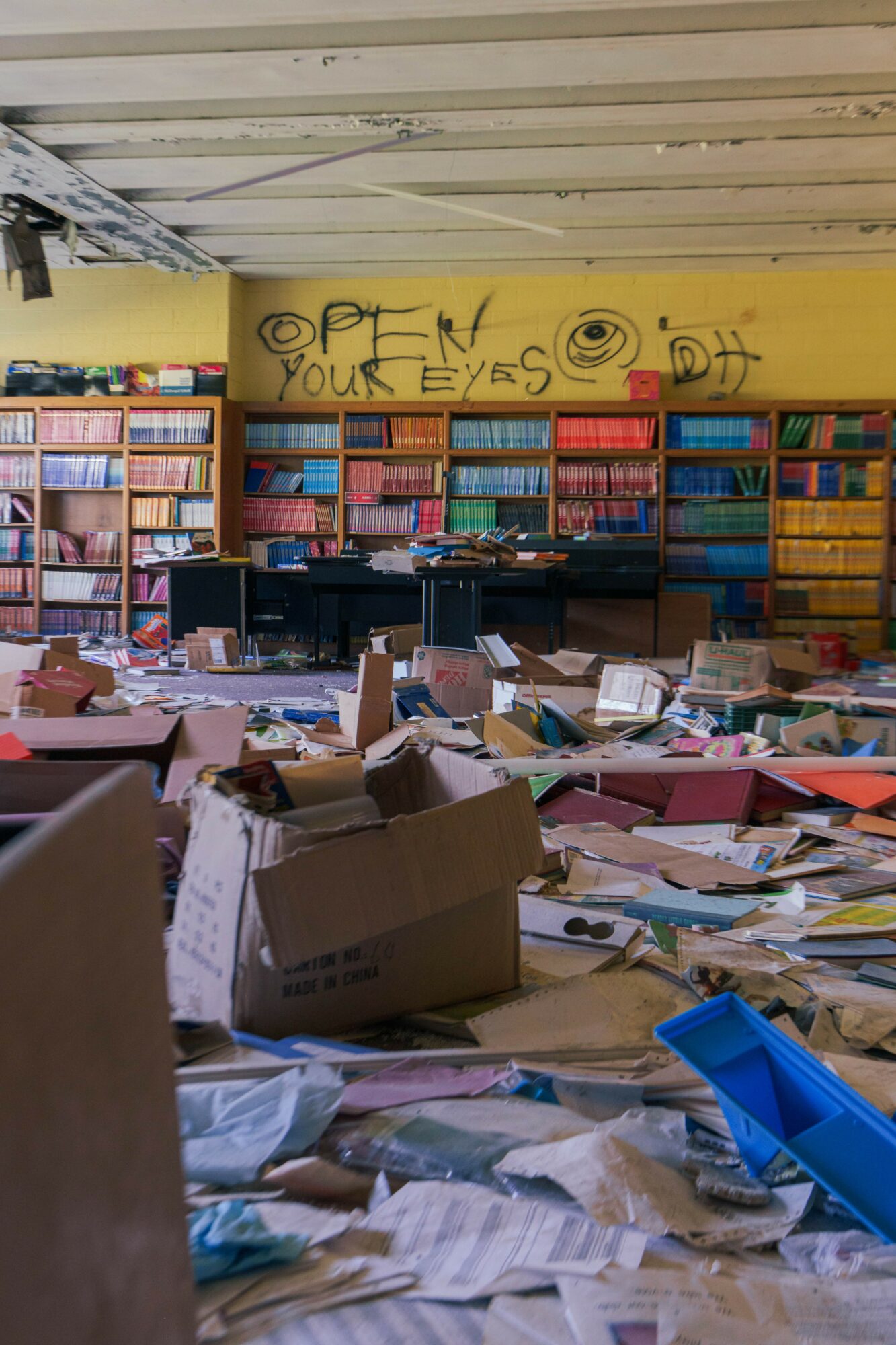 Room filled with scattered papers, boxes, and books, with colorful bookshelves and a yellow wall with graffiti in the background.