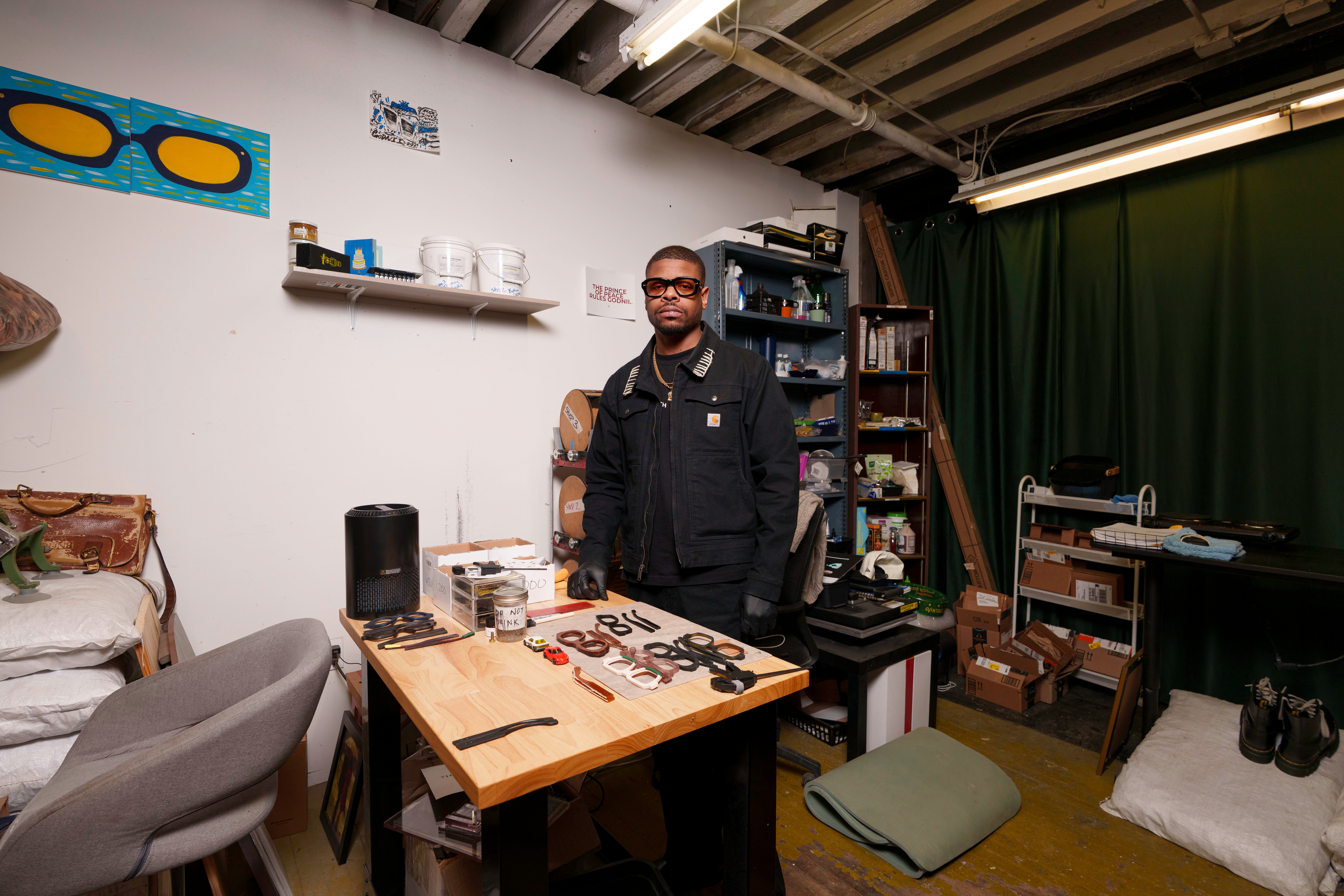 A man stands in a cluttered room with shelves, a table, and a bed, looking at the camera.