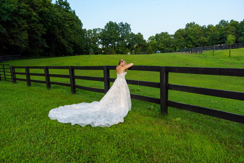 A wedding dress is draped over a black fence in a grassy field with trees in the background.