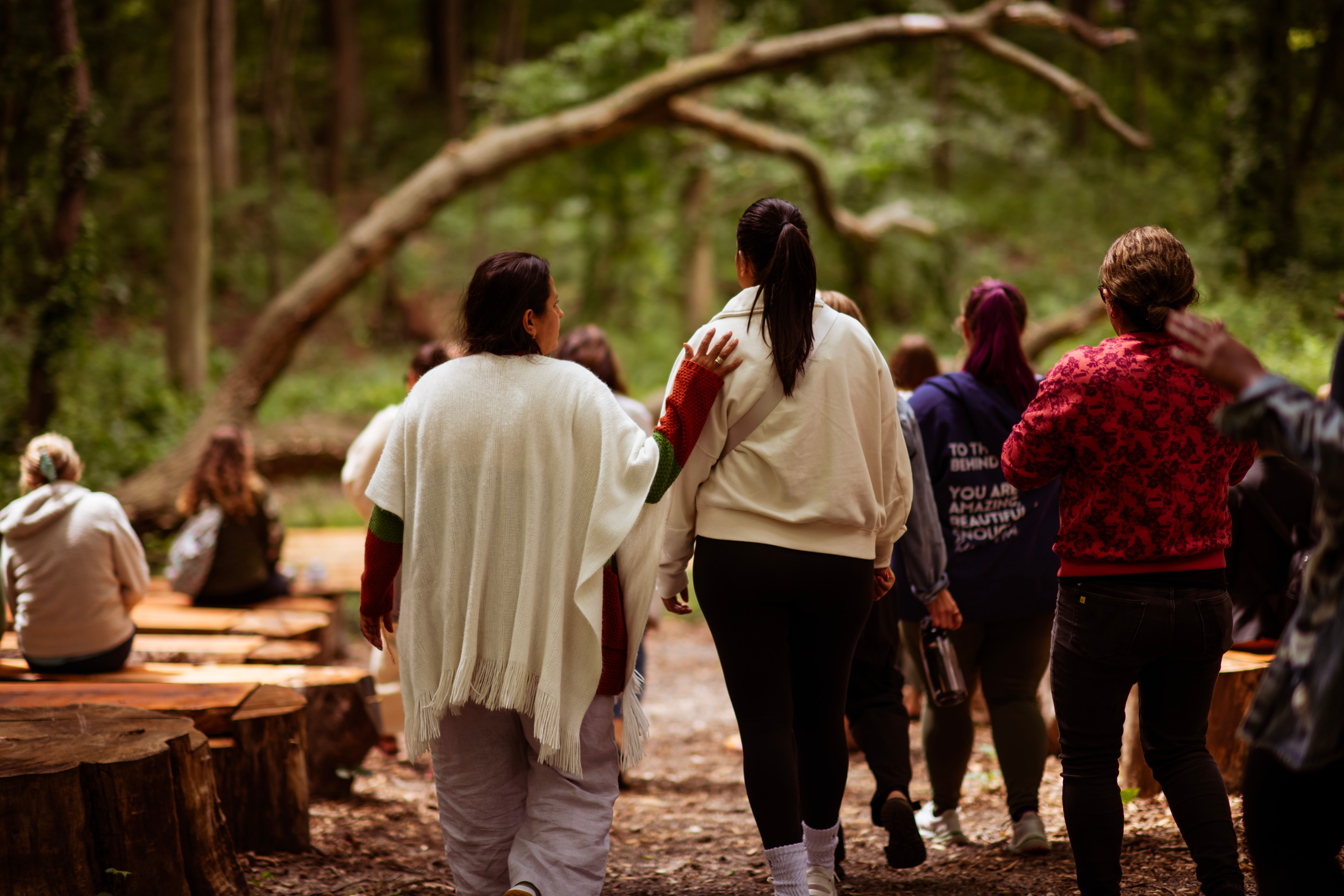 Group of people walking in a forest with trees and greenery, some sitting on logs, some standing and talking.