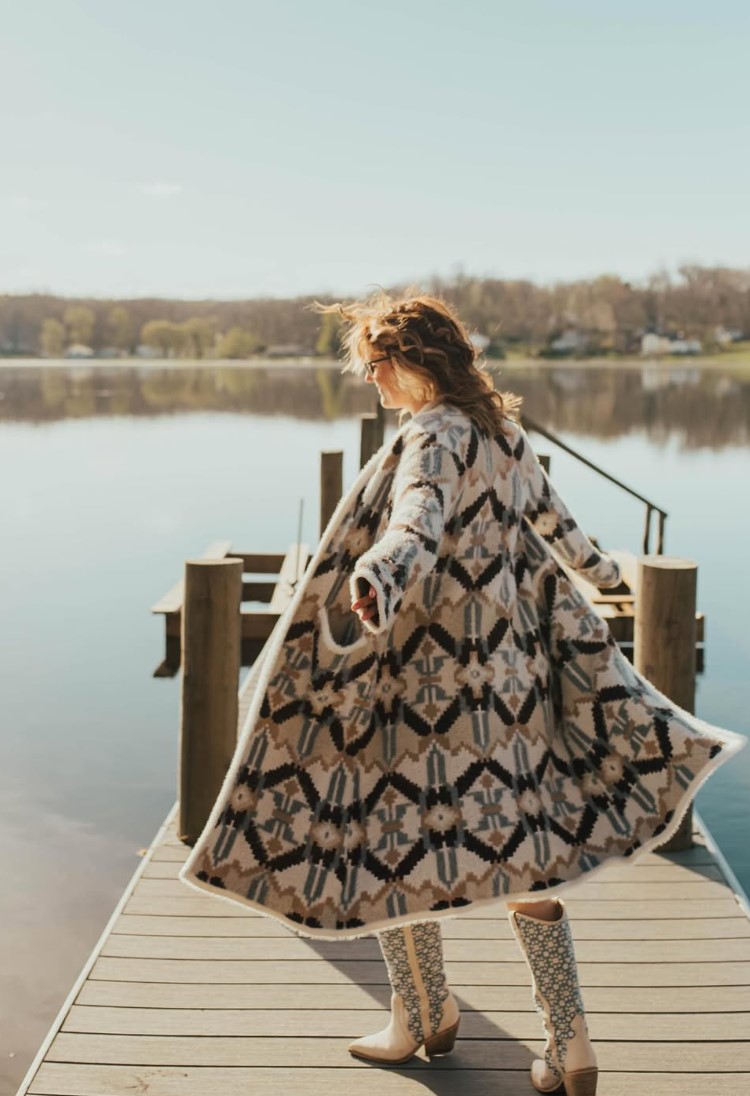 Person in patterned dress walking on a dock by a calm lake with trees in the background.