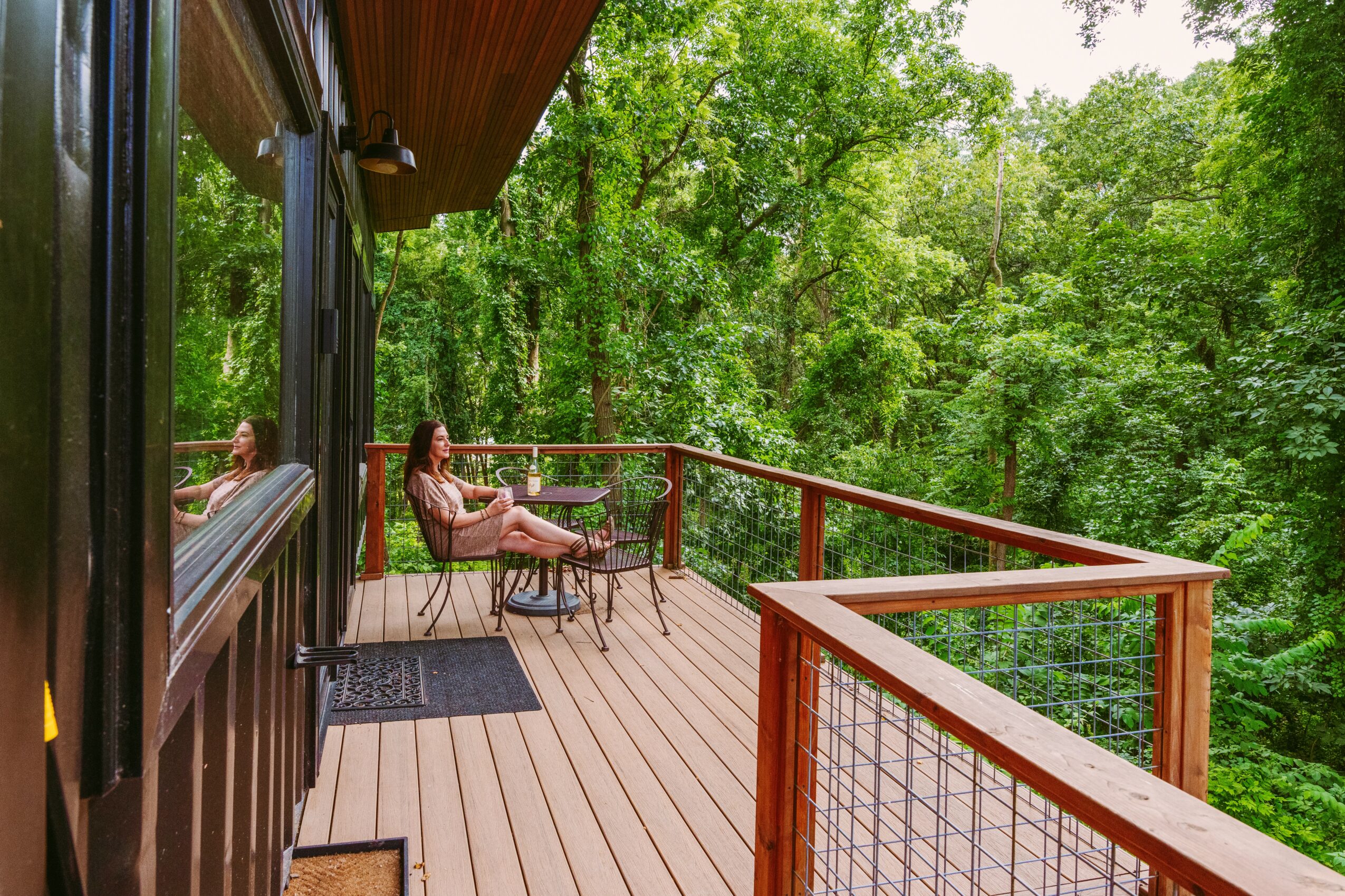Person sitting on a chair on a wooden balcony surrounded by green trees, with a railing and outdoor furniture.