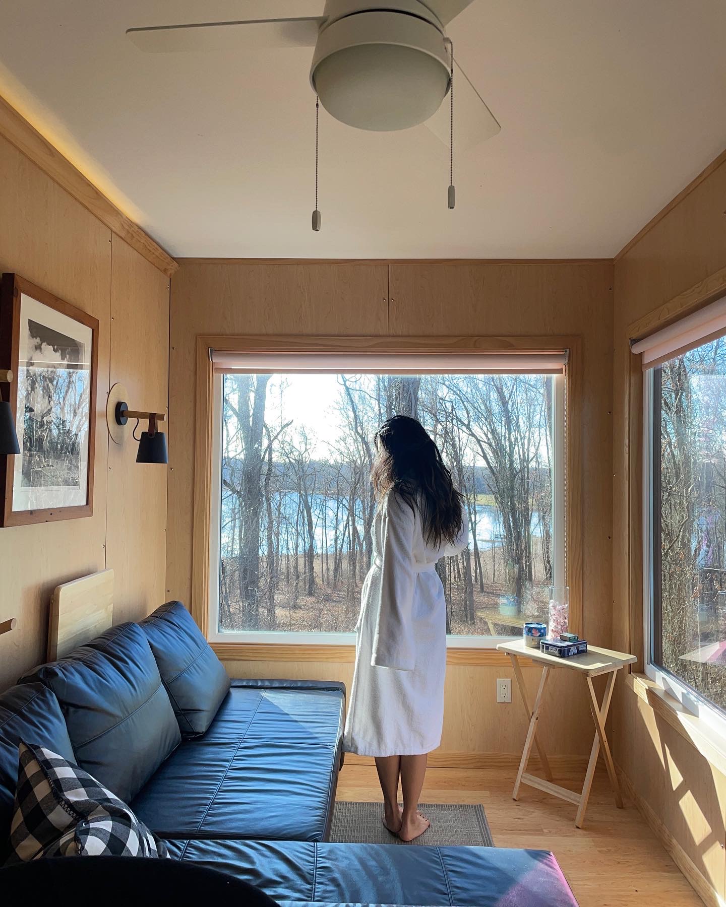 Woman in white dress standing by window overlooking trees and water, living room with blue sofa and wooden walls.