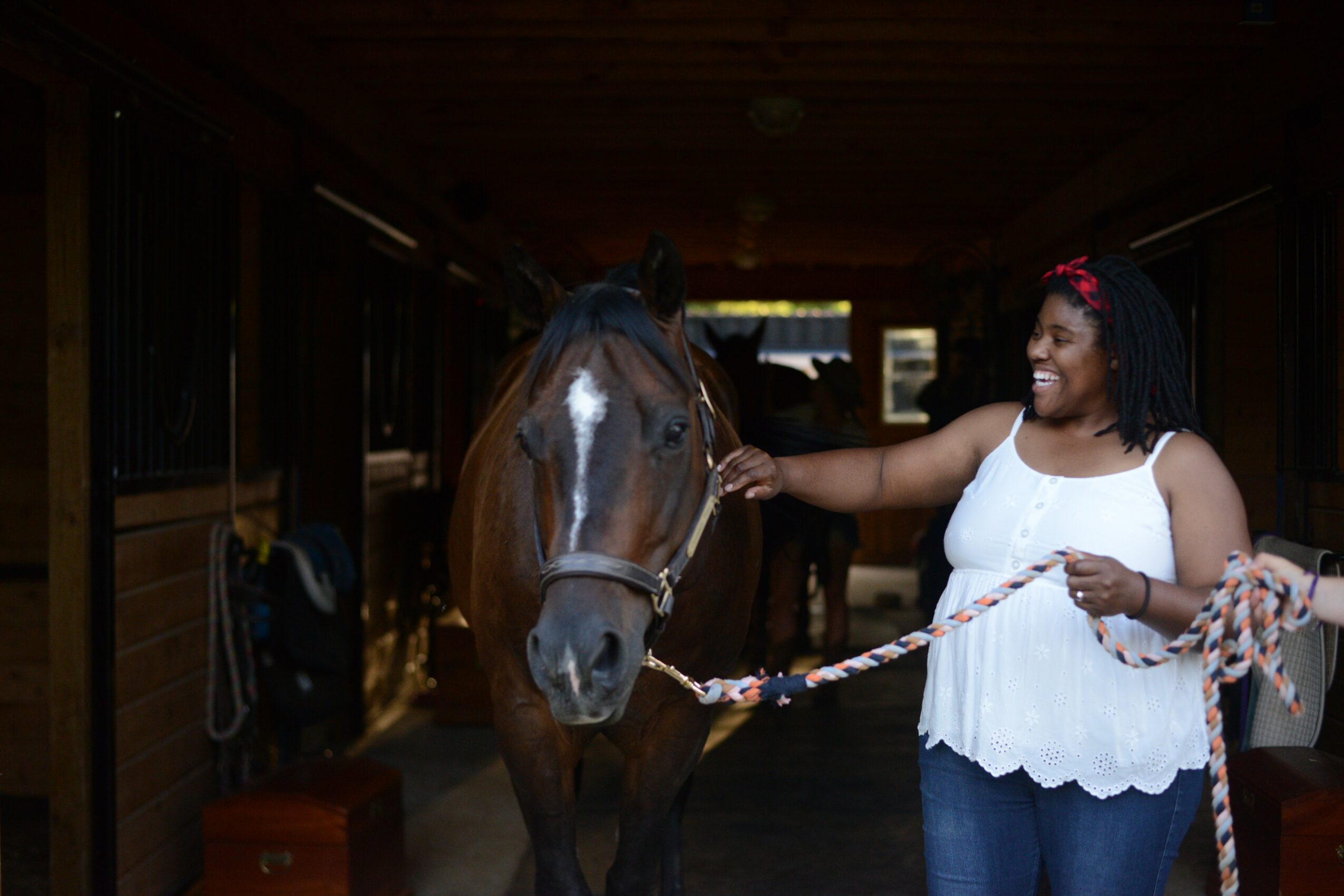 Smiling woman in white top petting a brown horse inside a stable.