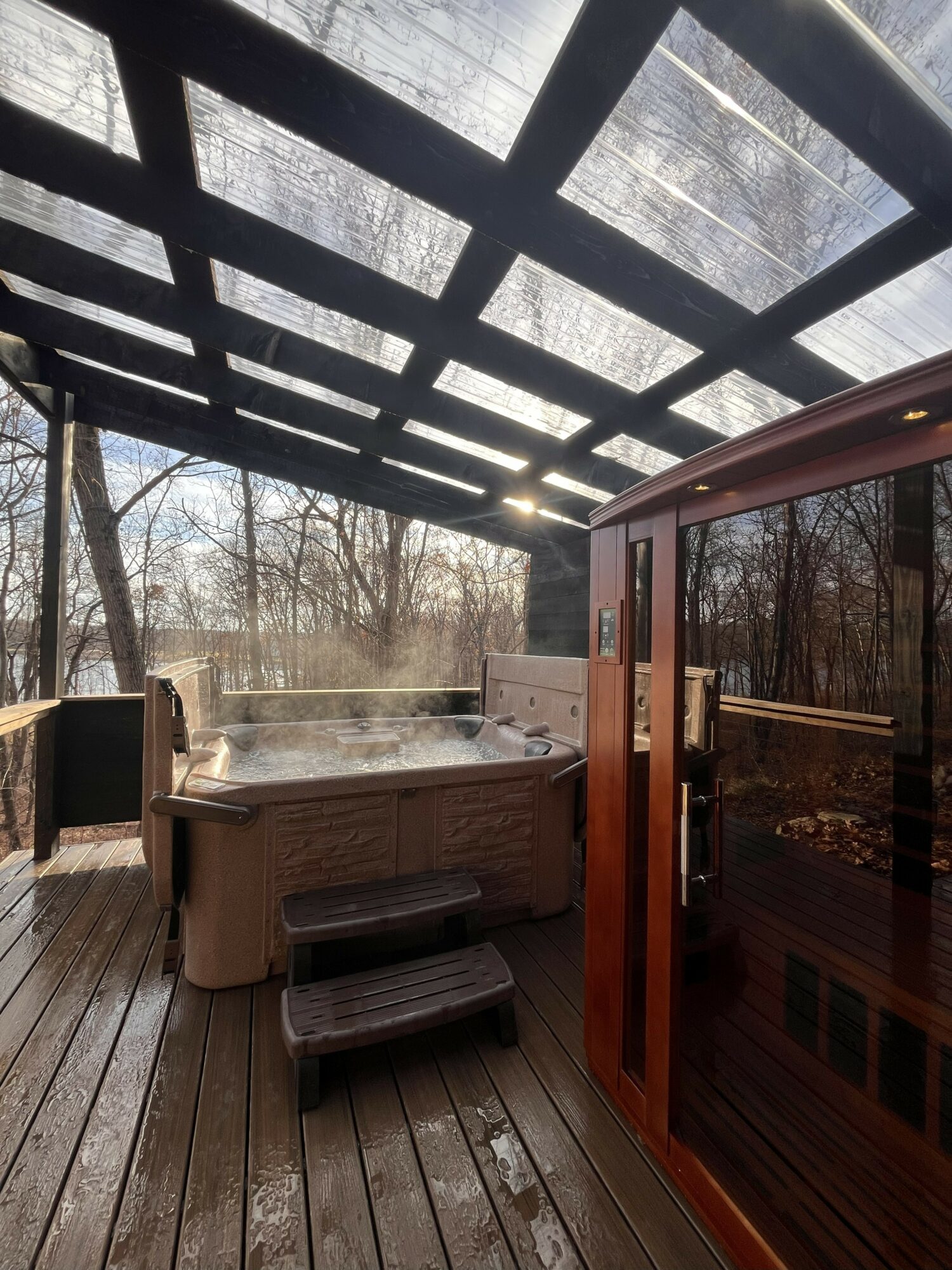 Hot tub on a wooden deck under a glass roof, surrounded by trees, with a wooden door nearby.